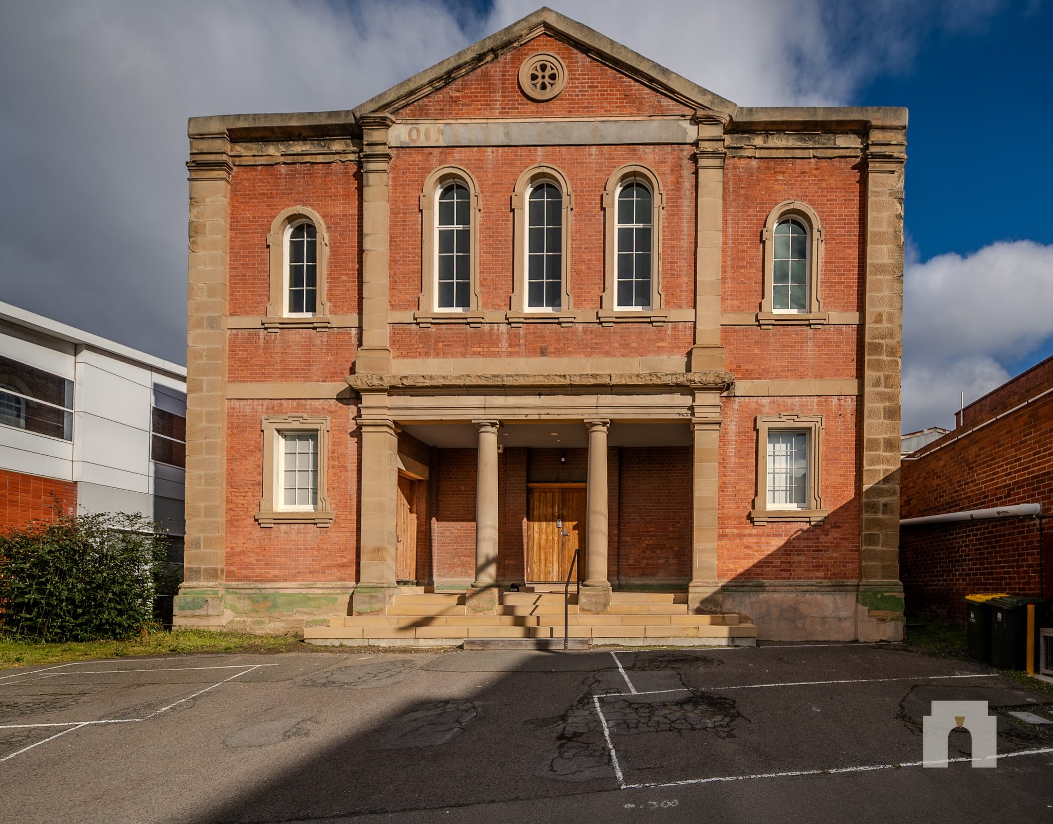 Two storey red brick building with sandstone trim, columned entry porch and car park at the front.