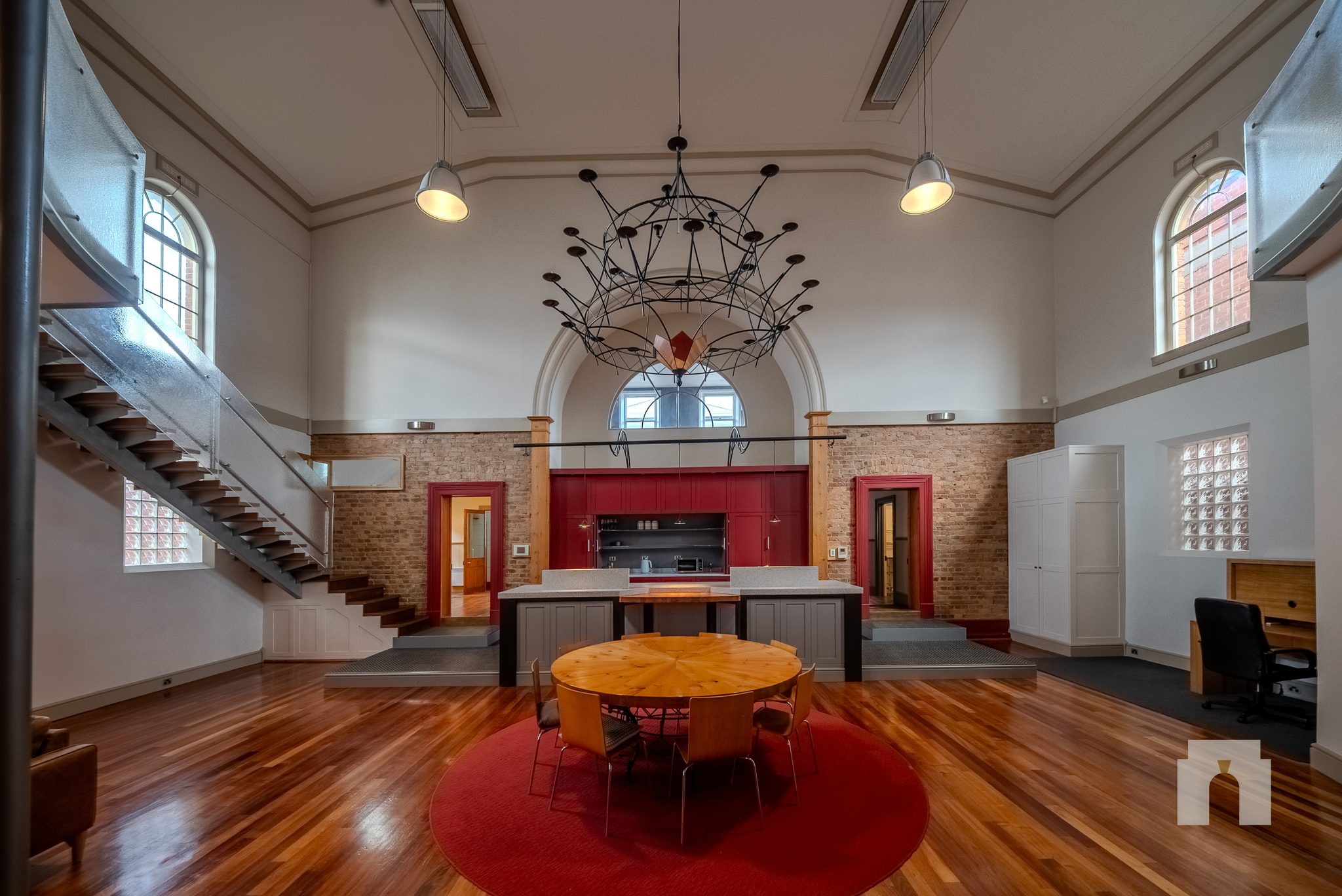 Open hall with timber floor, round table on a red rug, kitchen bench and stairs to a mezzanine.