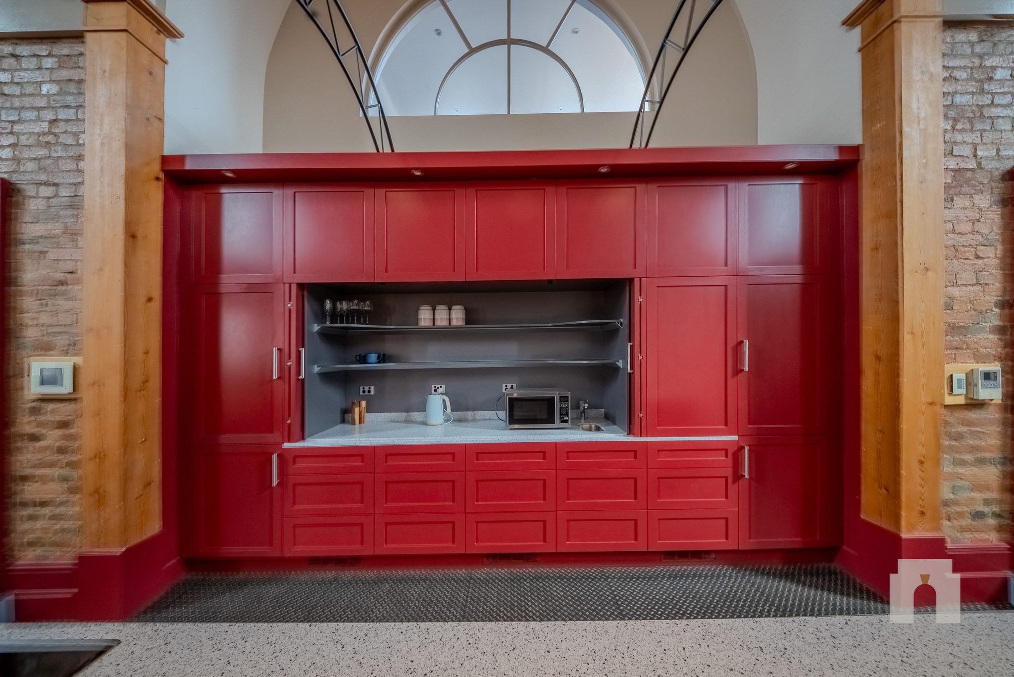 Red joinery kitchenette with open shelving, sink and microwave, set between timber posts and brick.