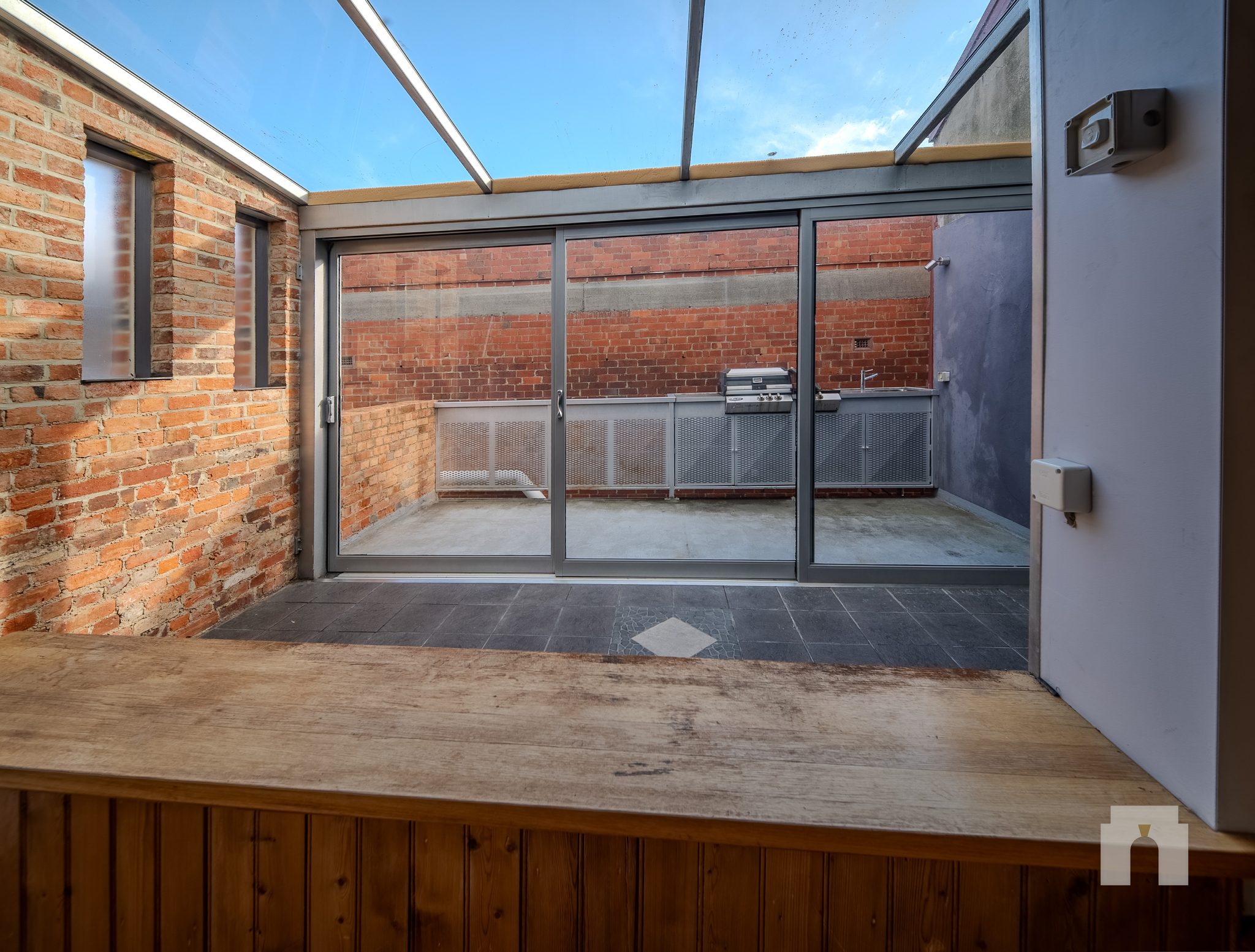 Glass roofed space with brick walls, sliding doors to a paved terrace with barbecue and sink.