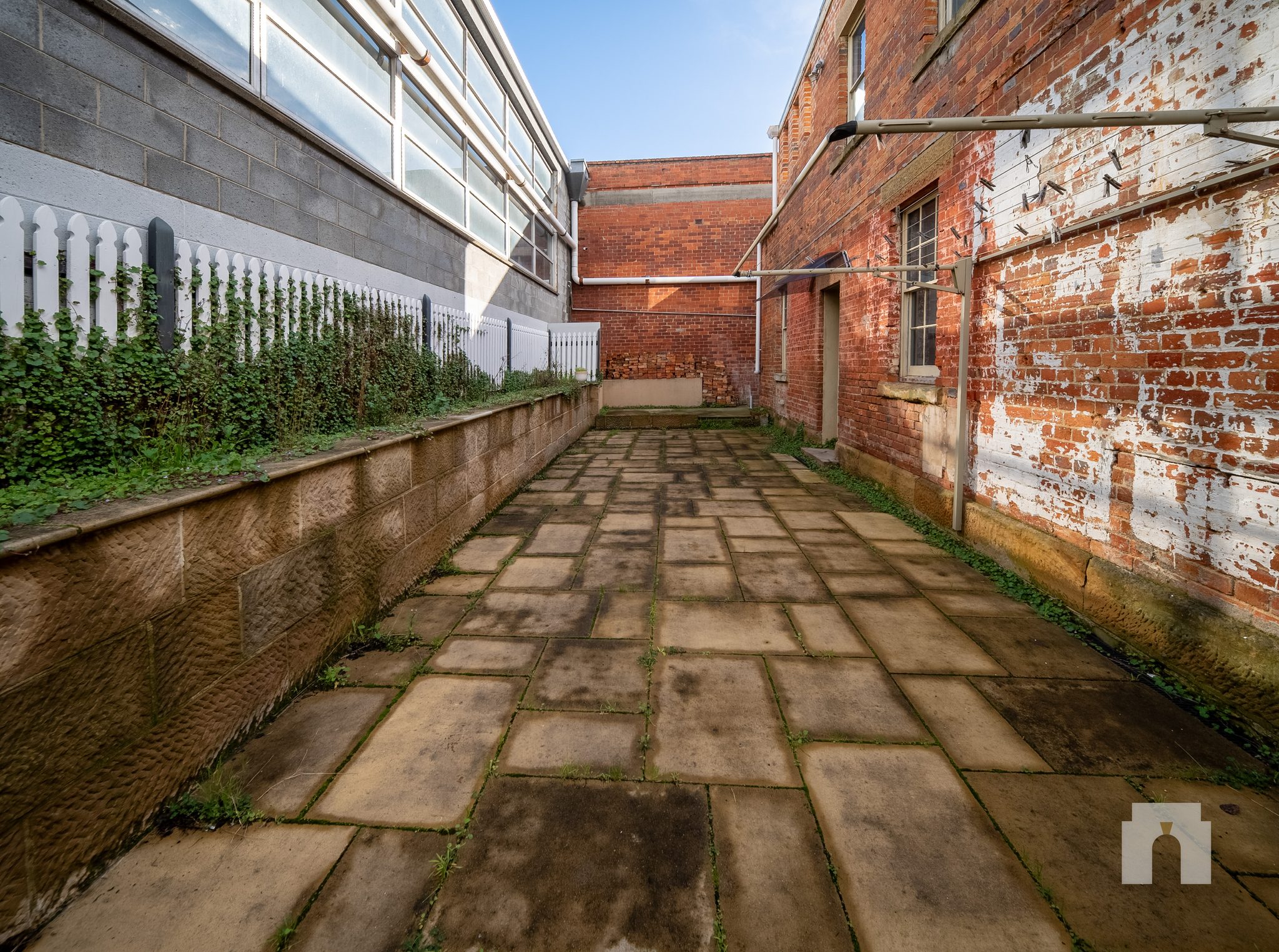 Long paved side courtyard between a brick wall and a rendered wall topped with picket fencing.