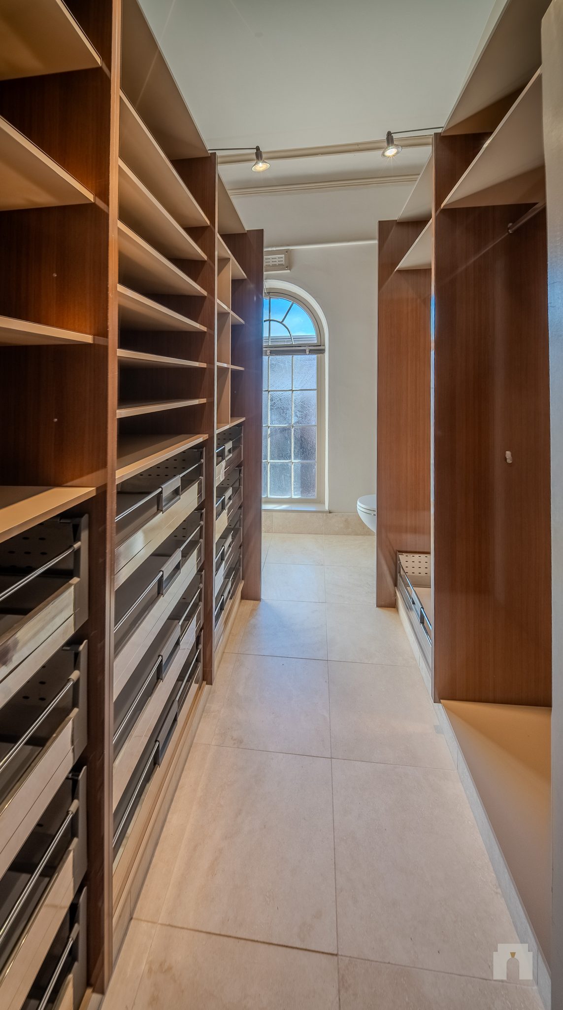 Narrow tiled room lined with timber shelving and pull out drawers, arched window at the end.