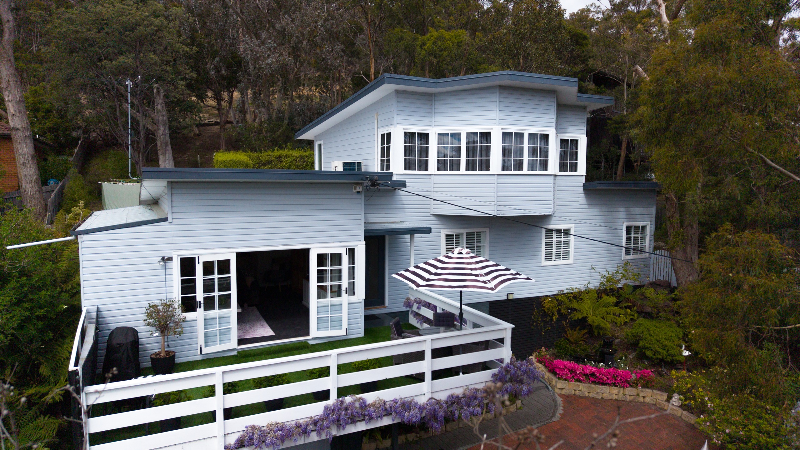 Aerial view of the weatherboard house set into the hillside among gum trees, with a white-railed deck and a striped umbrella, and wisteria and pink azaleas below.