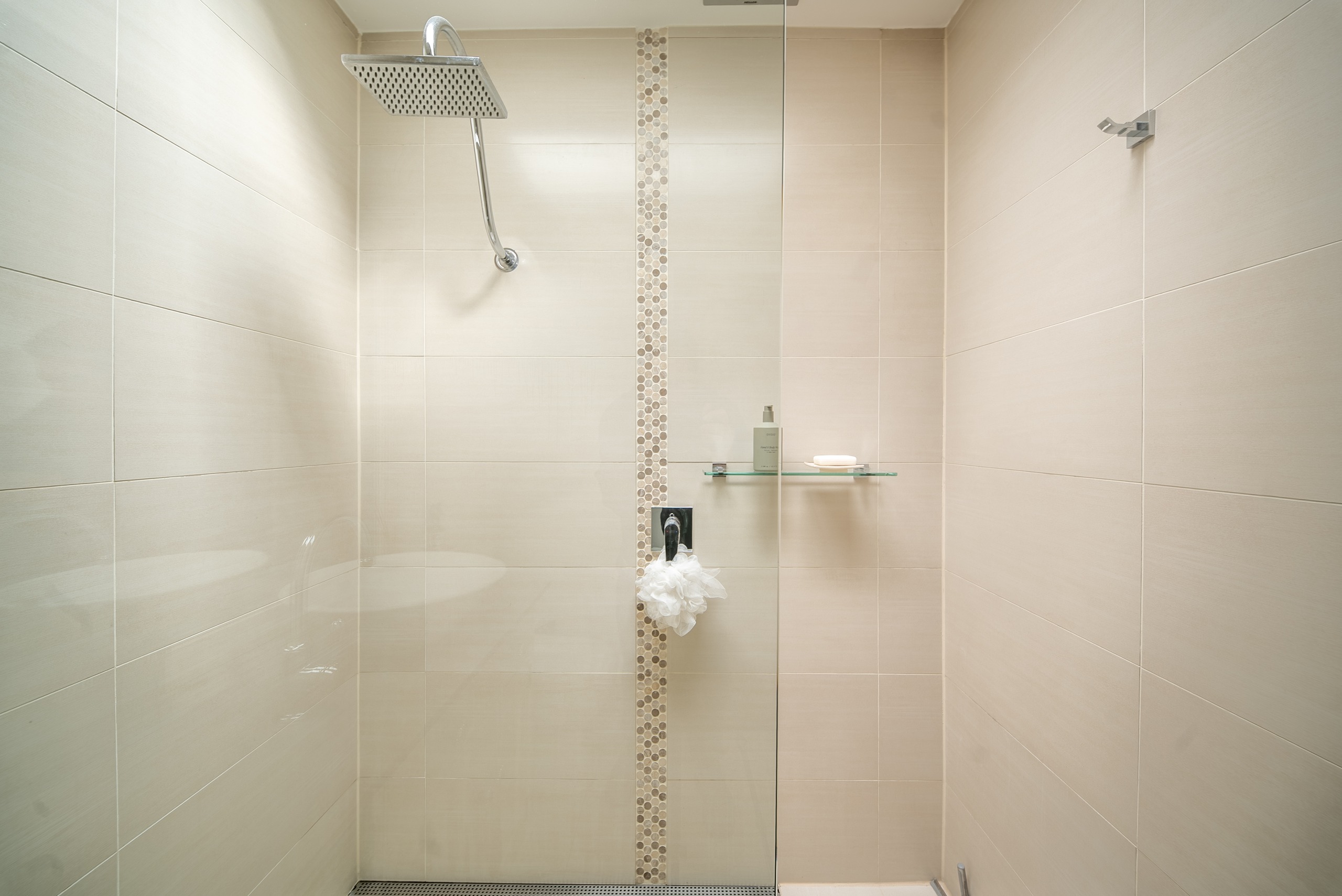 The walk-in shower with a rain showerhead, large cream tiles and a glass shelf.