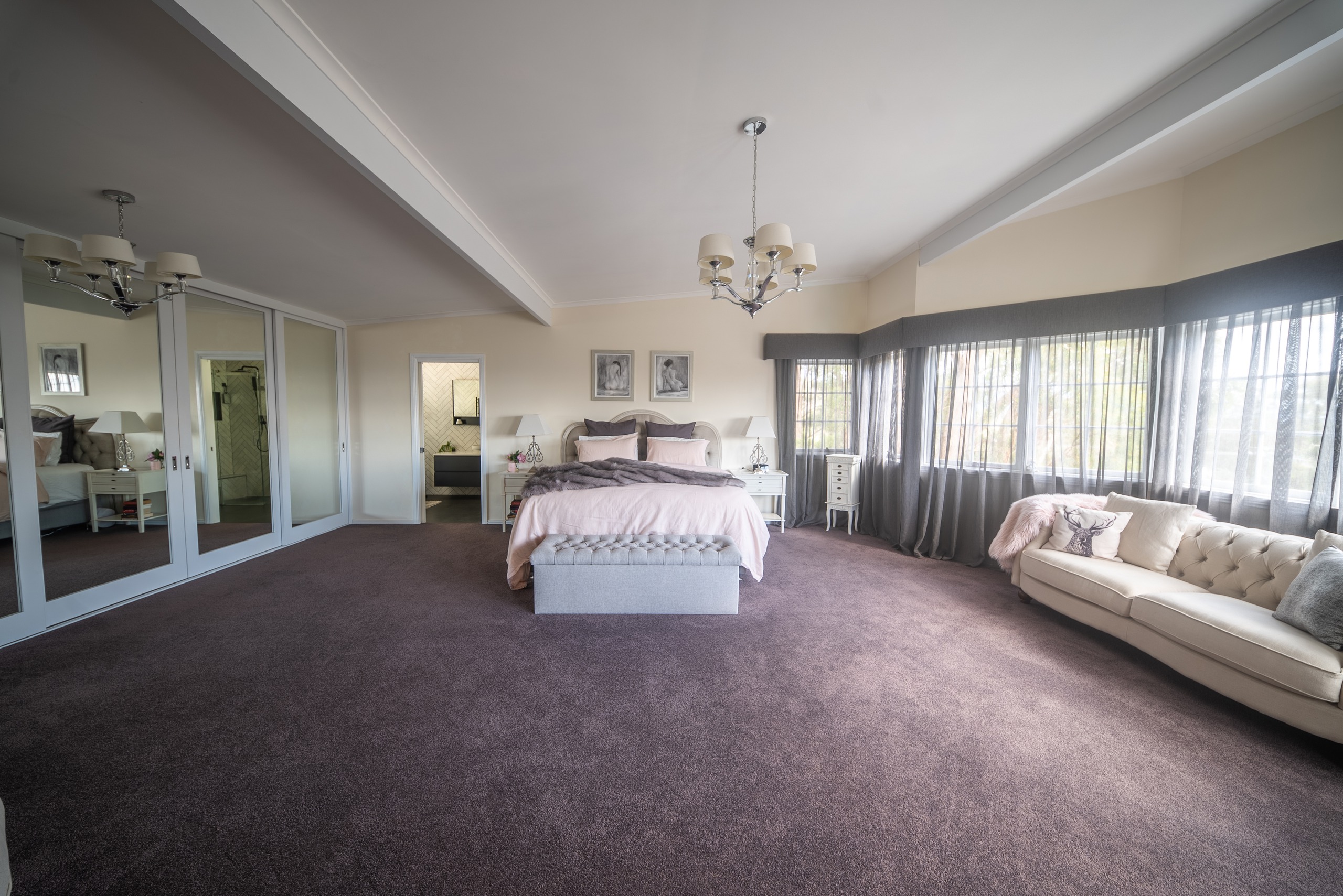 A large carpeted bedroom with a chandelier, mirrored wardrobe doors, a bay of windows and a cream sofa.