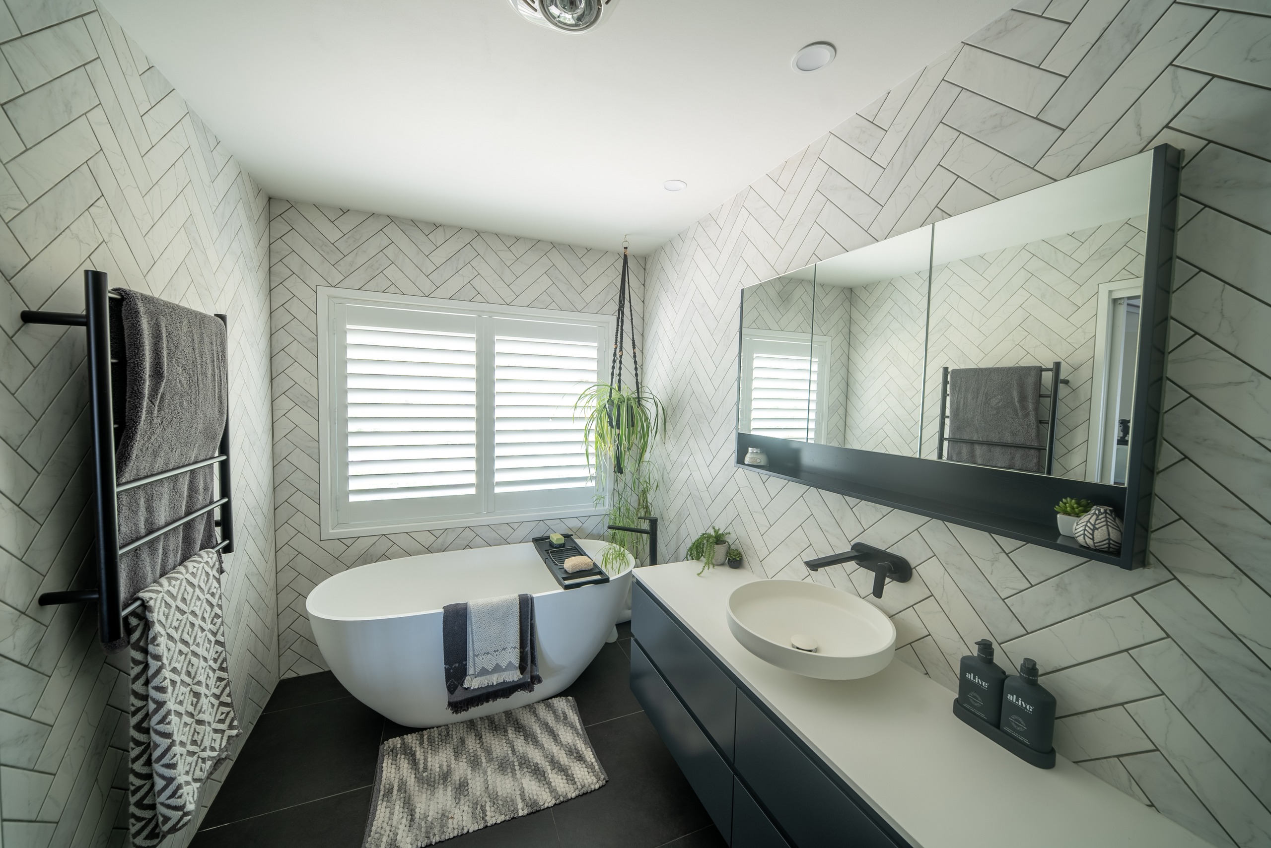 A bathroom with herringbone tiles, a freestanding bath, a black towel rail and a long vanity with a mirrored cabinet.