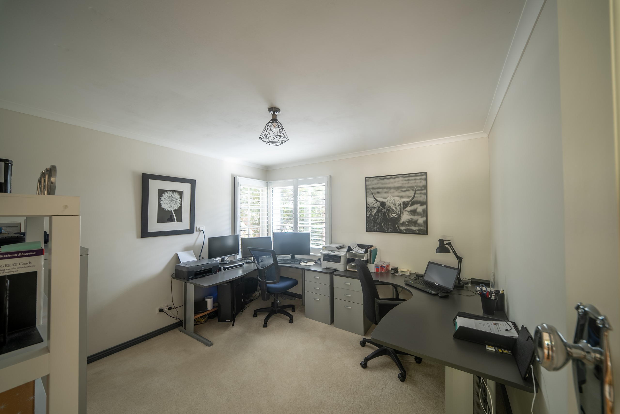 A study with two desks, computer monitors and office chairs, lit by a window with plantation shutters.