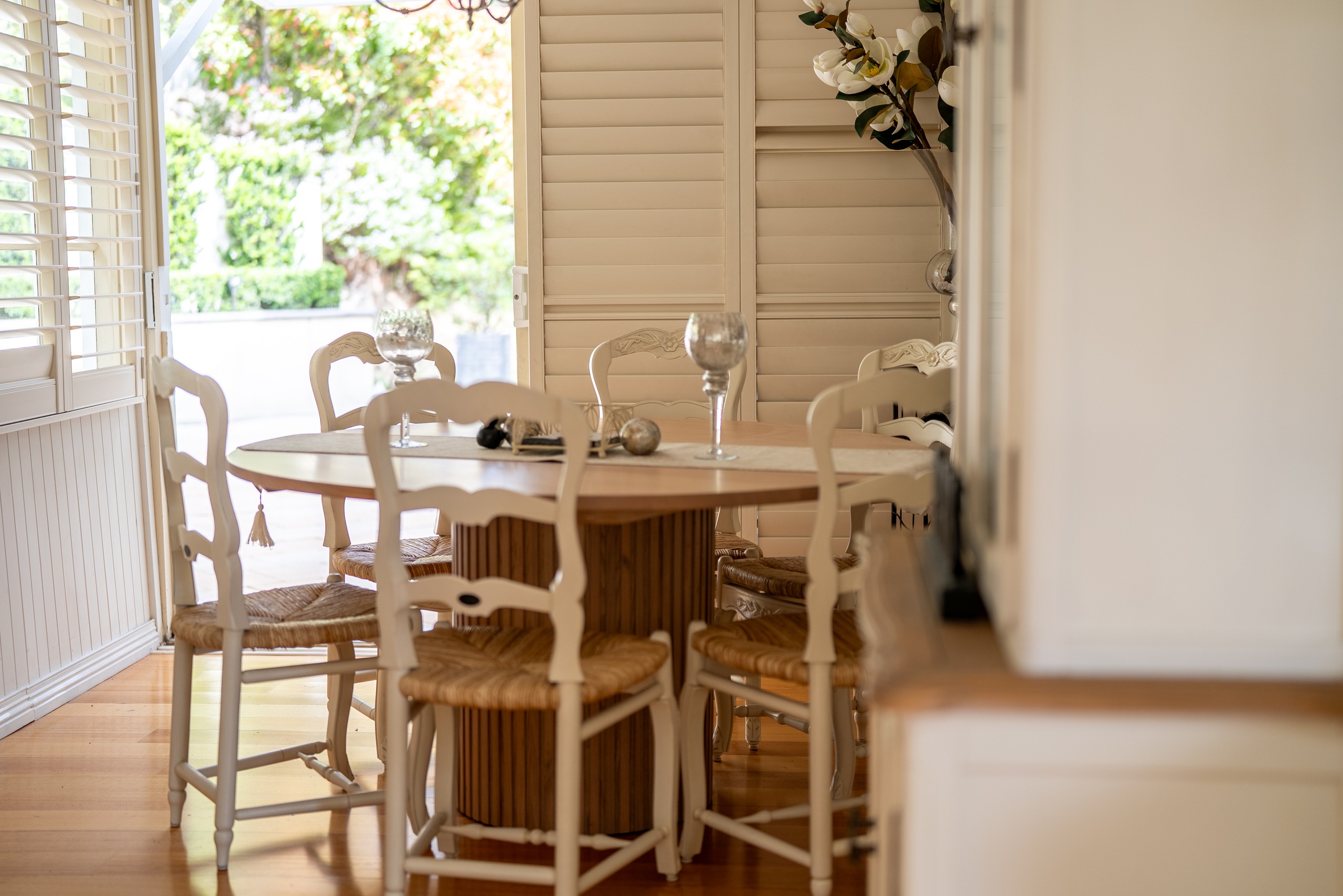The dining table and chairs by a window with plantation shutters, looking out to the garden.