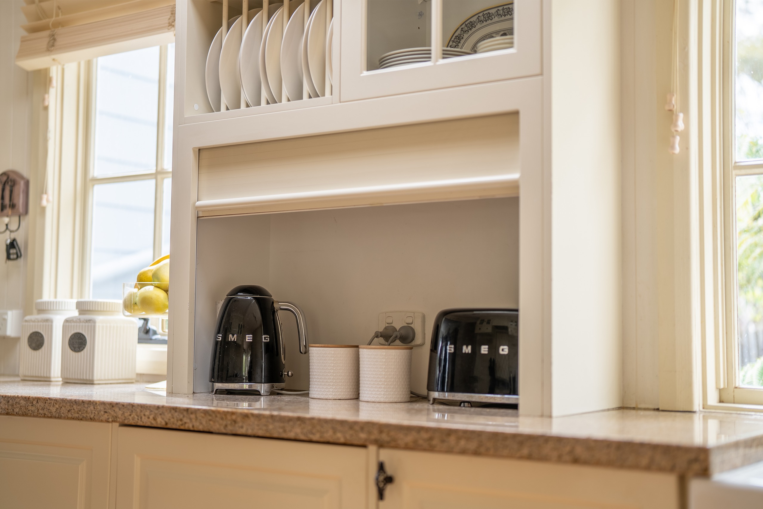 A kitchen nook with a kettle and toaster under a glass-fronted cabinet and plate rack.