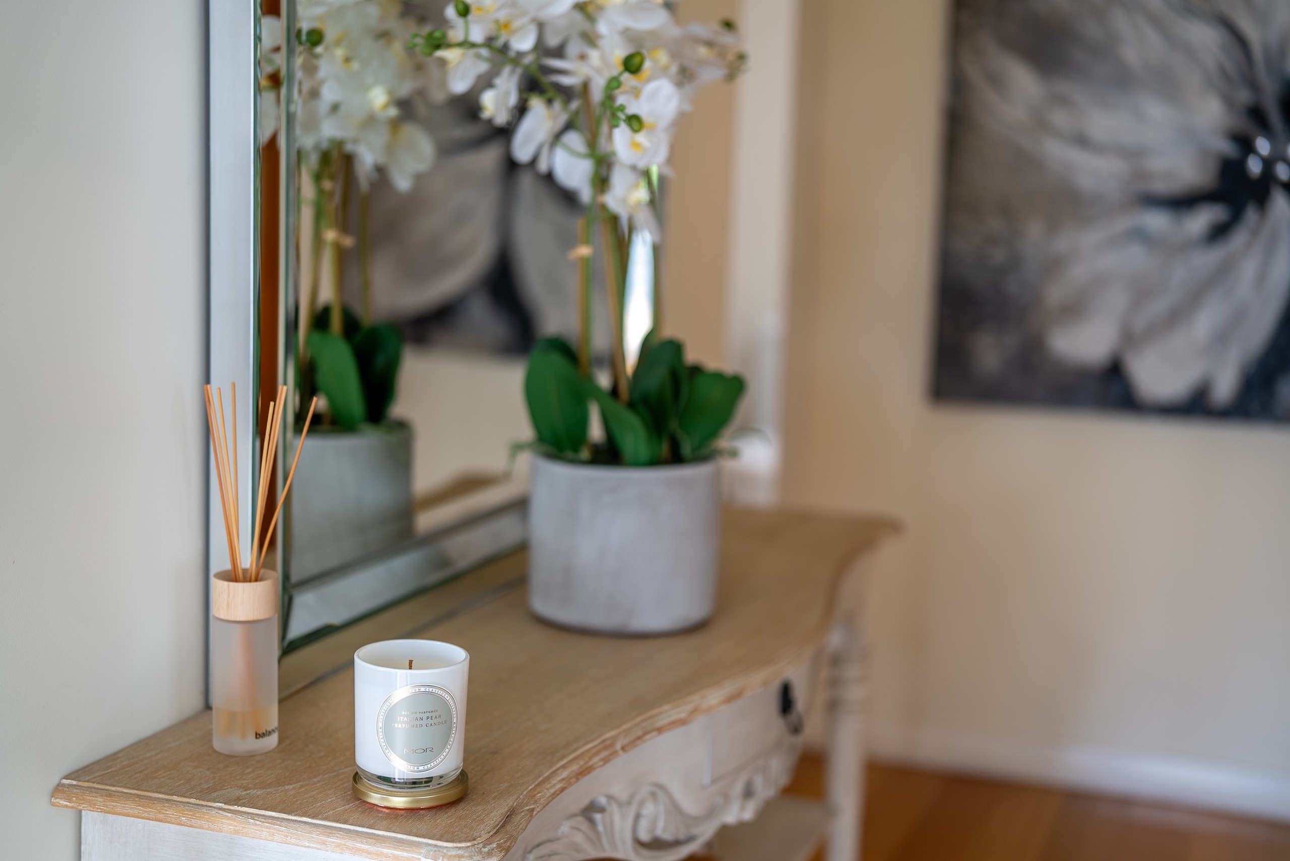 A console table with an orchid, a candle and a reed diffuser in front of a mirror.