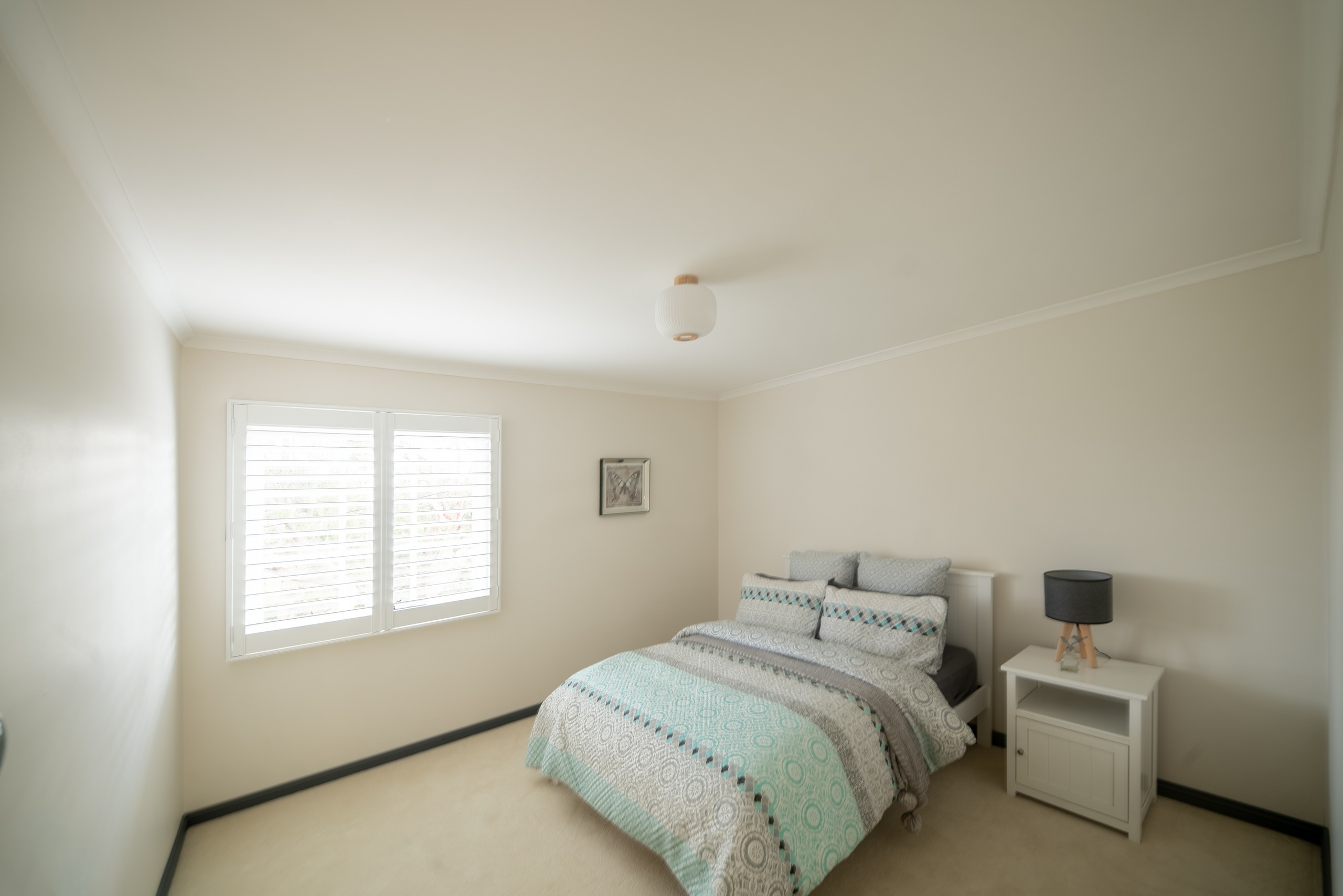 A carpeted bedroom with a queen bed, a bedside table and lamp, and a window with plantation shutters.
