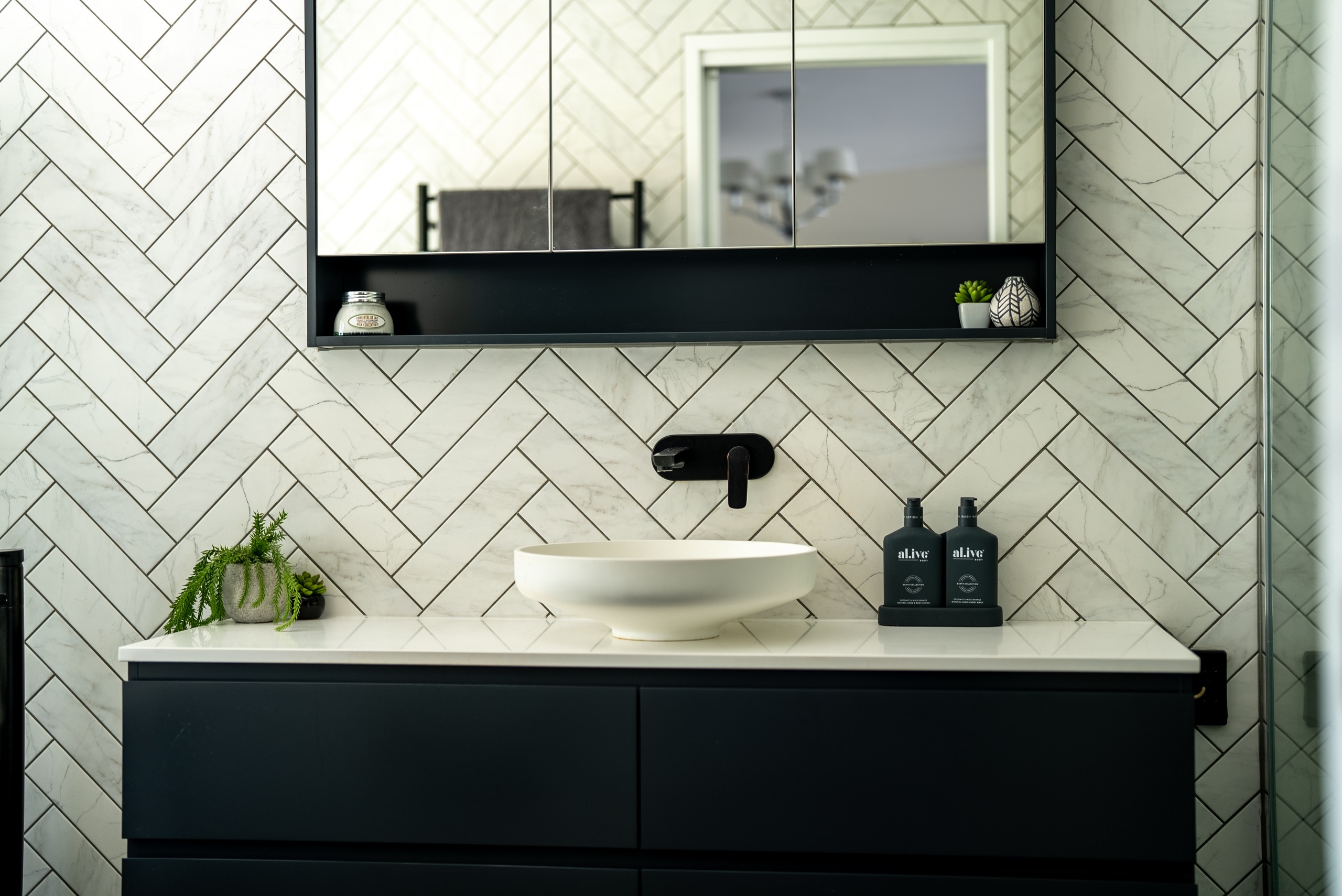 A black vanity with a round vessel basin and a black wall mixer against white herringbone tiles.