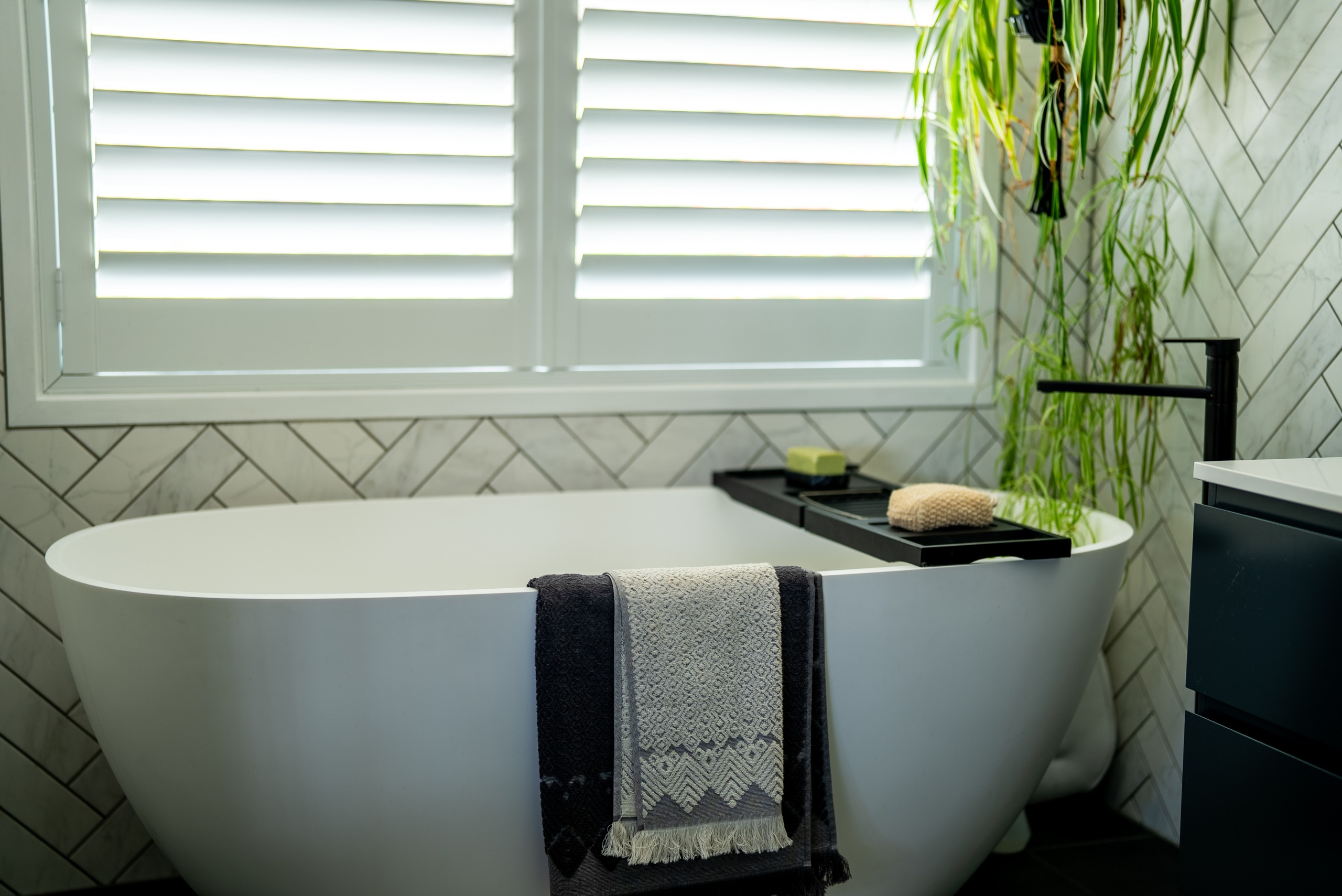 A freestanding bath under a window with plantation shutters, with a trailing plant beside it.