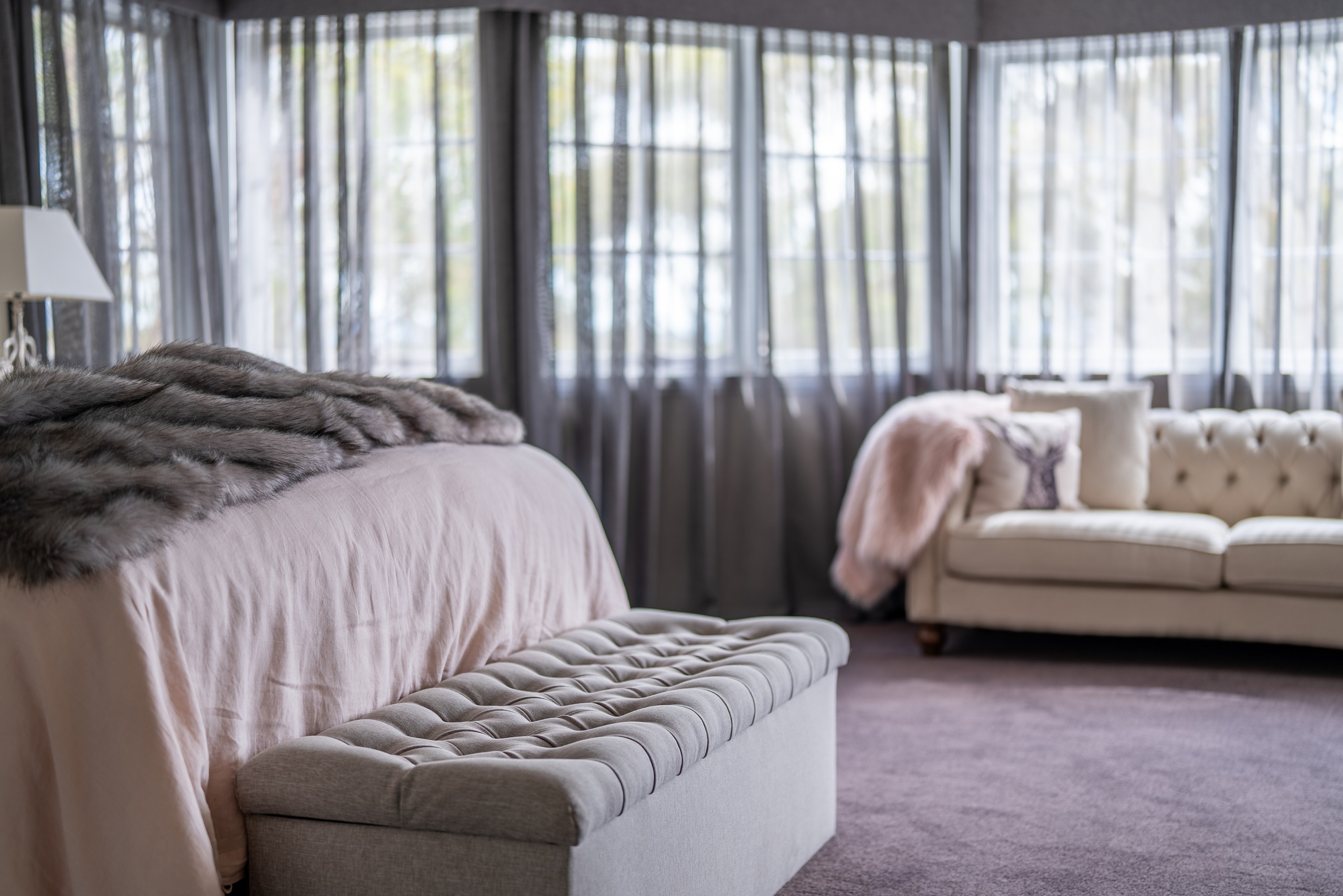 The main bedroom bay window with sheer curtains, a cream tufted sofa and the end of the bed.