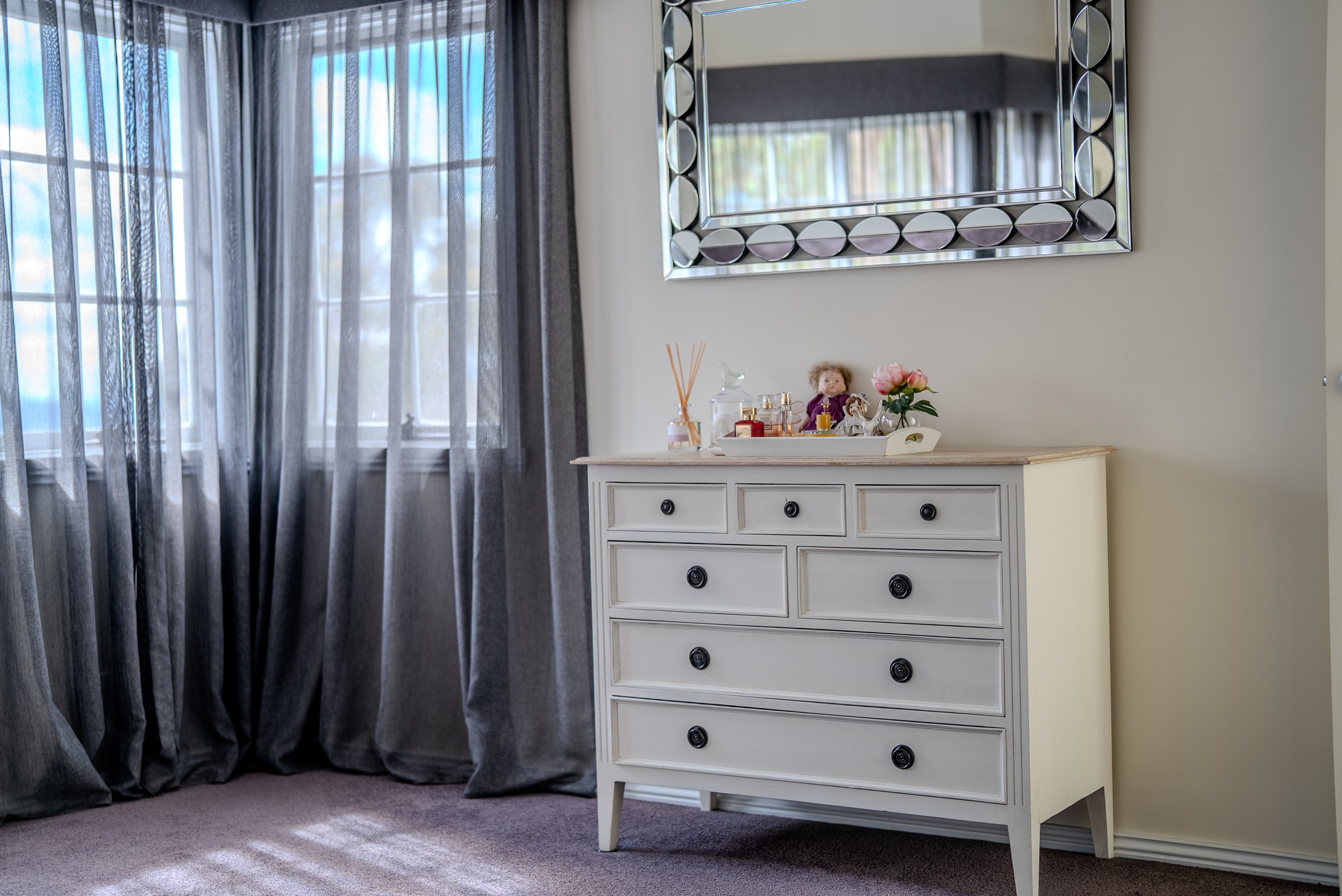 A white chest of drawers beneath a mirror, between windows with sheer grey curtains.