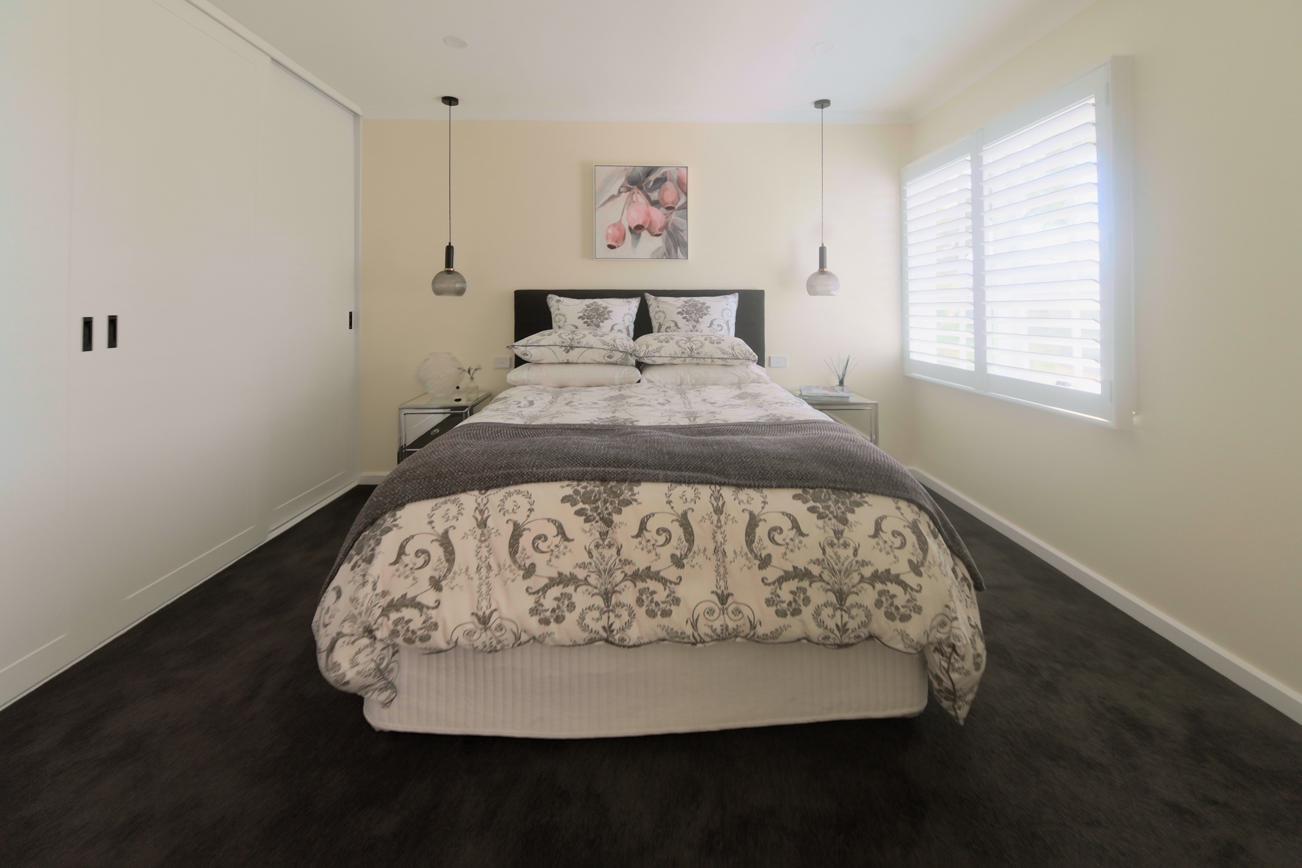 A bedroom with built-in wardrobes, a queen bed with pendant lights either side and a botanical painting above it, dark carpet in front of the bed, and a window with plantation shutters.