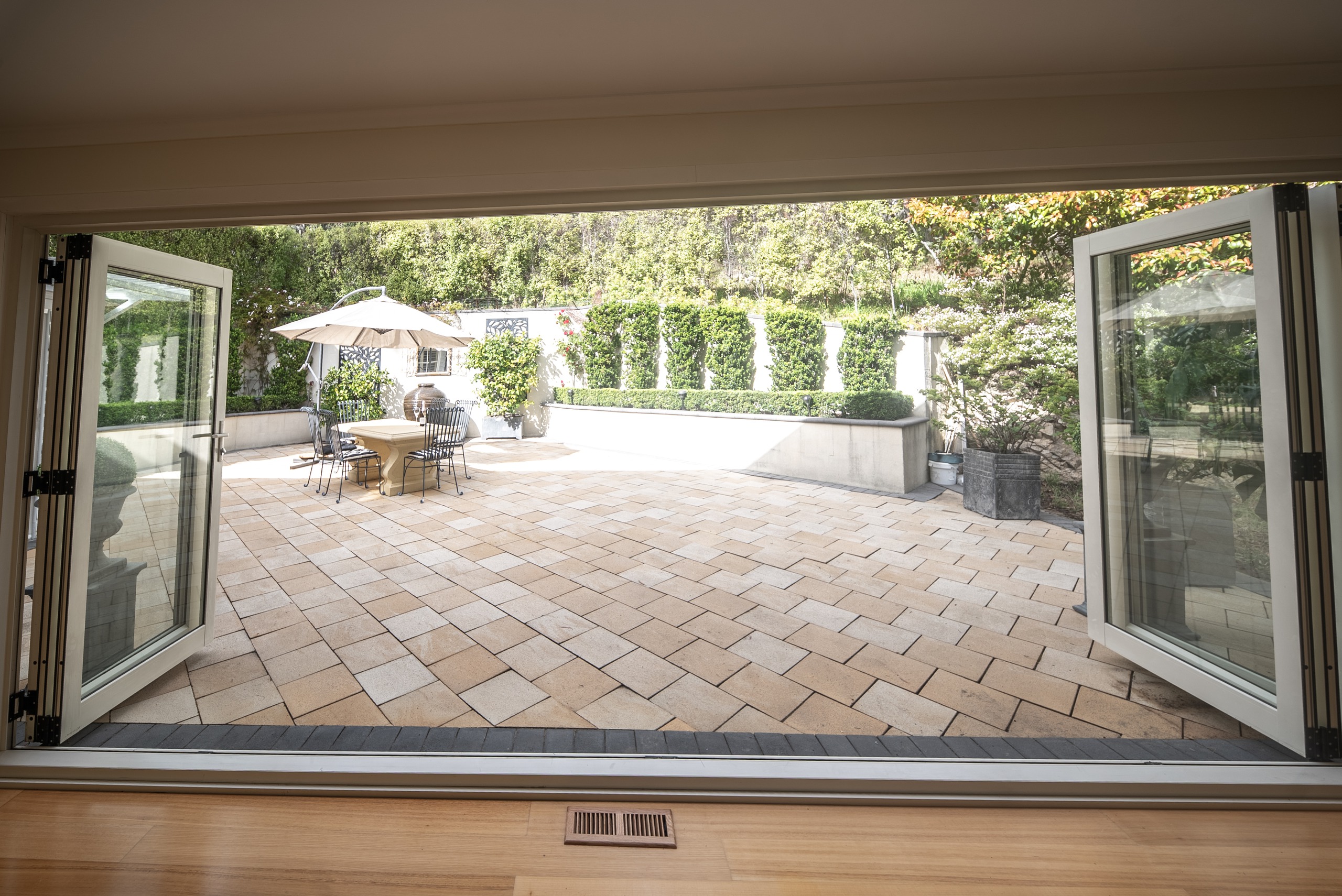 Looking out through open bifold doors from inside the house to the paved courtyard, with an outdoor table and umbrella and clipped hedges beyond.