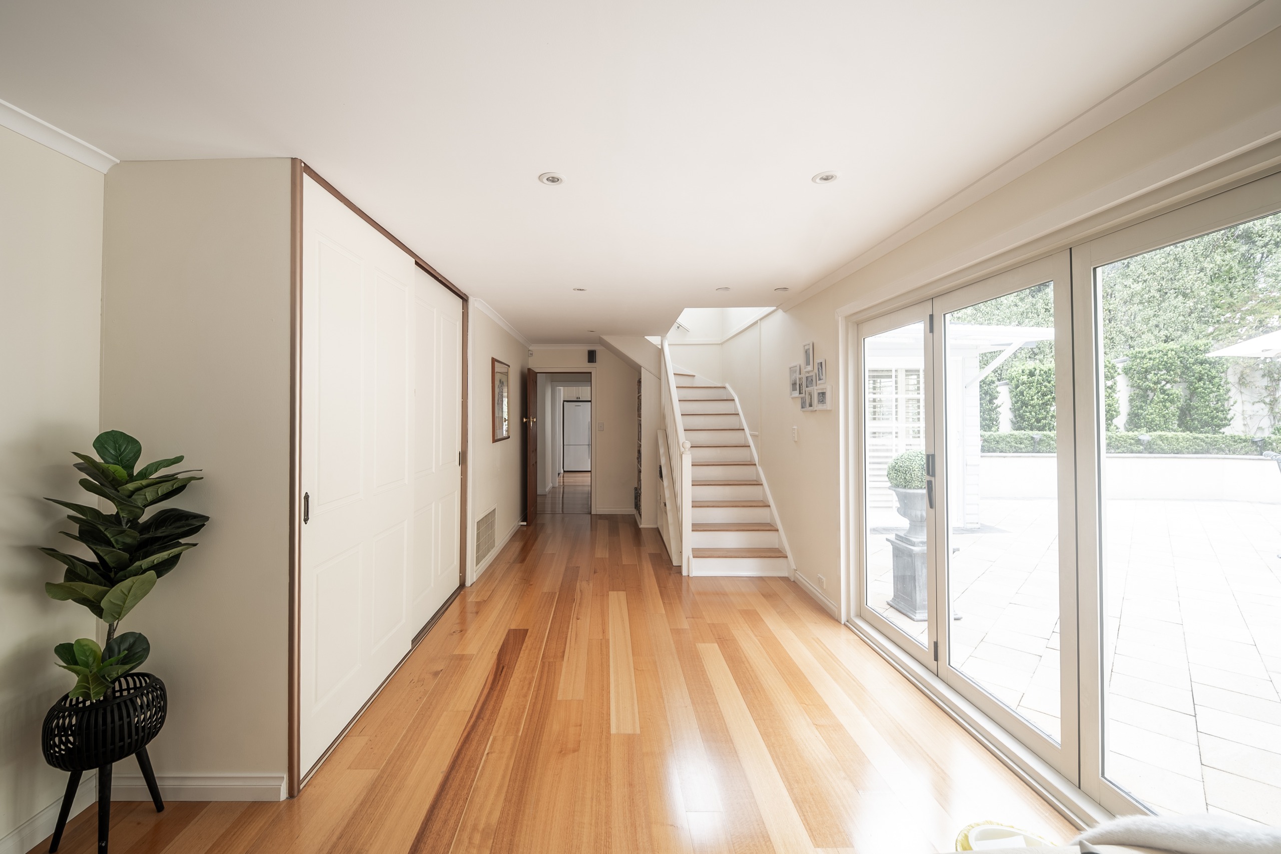 A hallway with timber floors and built-in cupboards, glass sliding doors along one side to the paved courtyard, and a staircase at the far end.