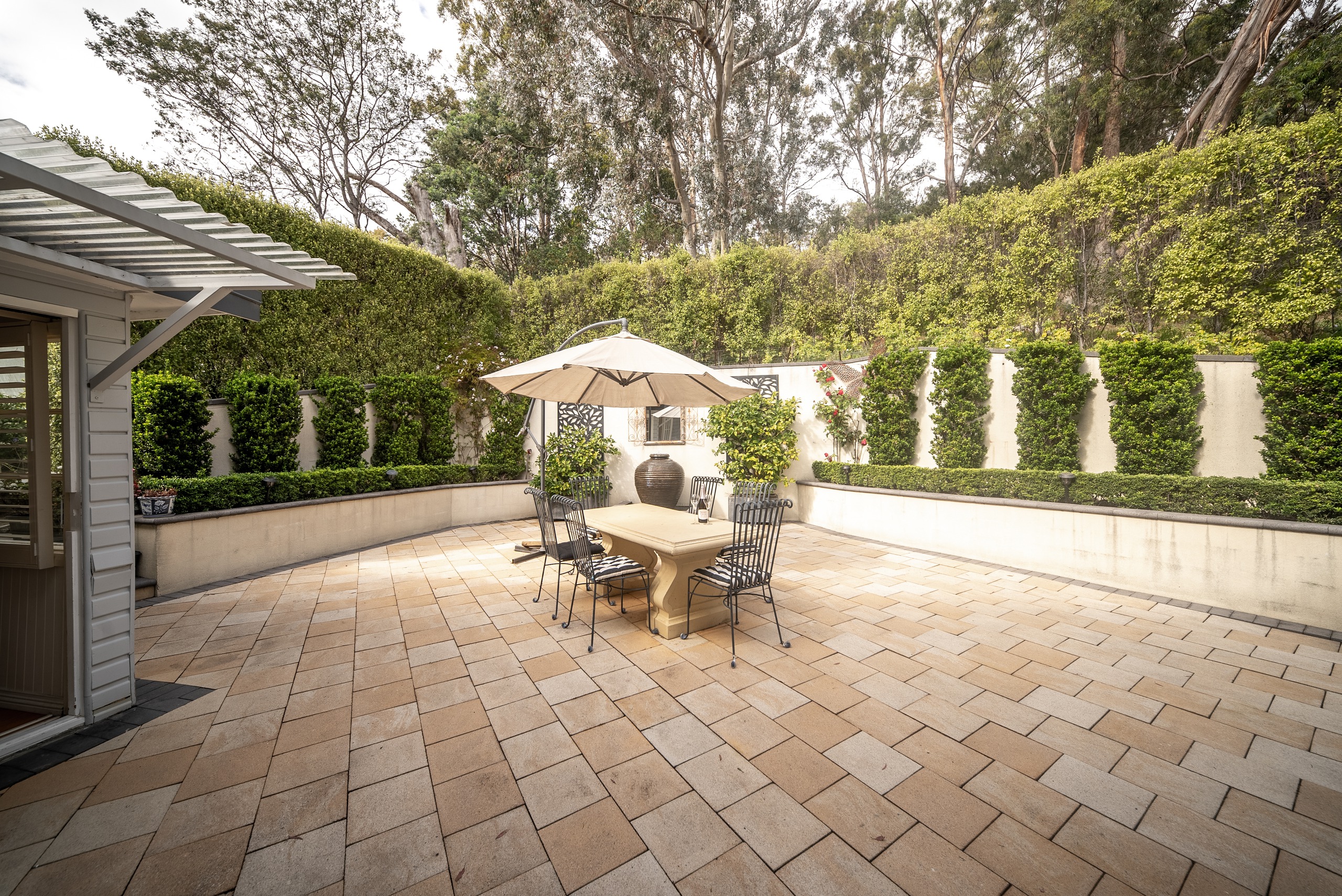 The paved courtyard with an outdoor table and umbrella, enclosed by high hedges and trees.