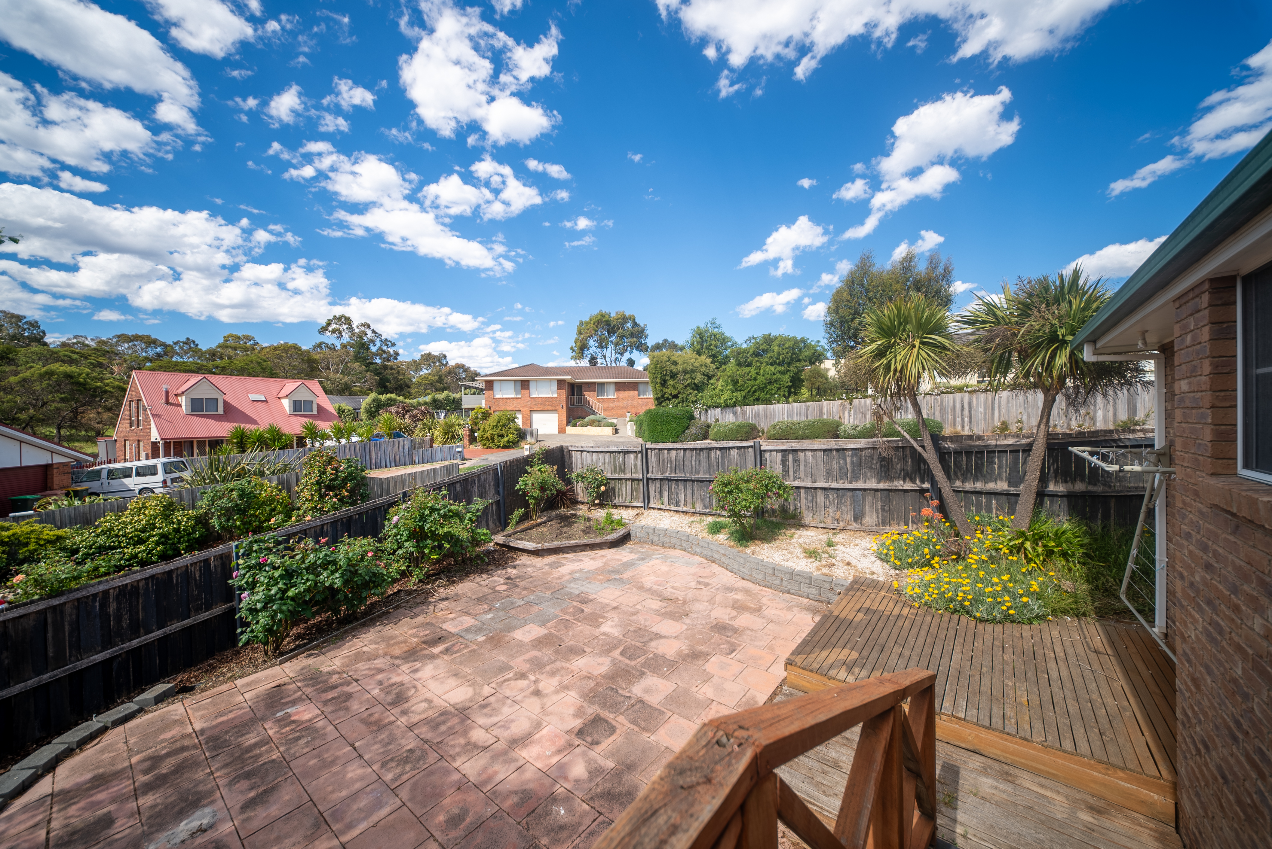 Backyard seen from the house, with paving, a timber deck and garden beds along the fence.