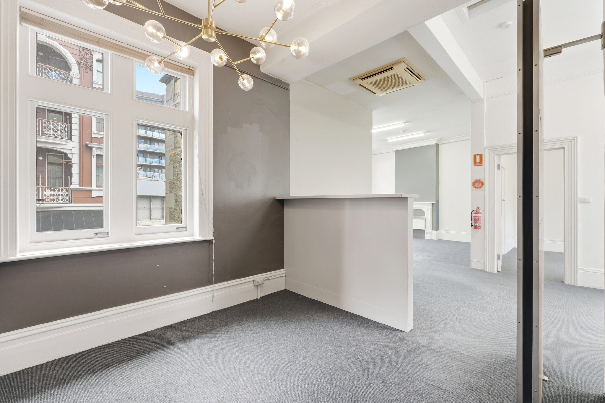 Empty carpeted room with a reception counter, sash windows to the street and a ceiling air conditioning unit.