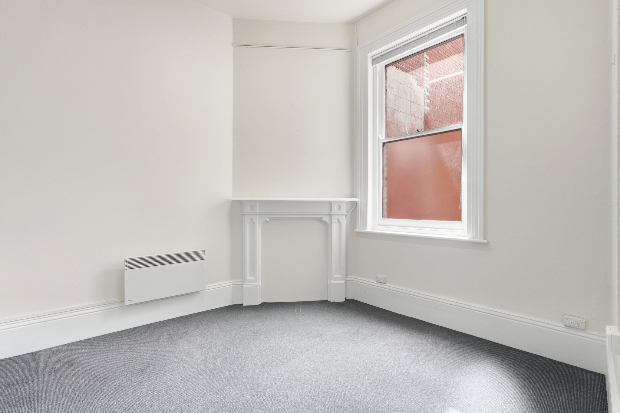 Small carpeted room with a painted mantelpiece, a wall panel heater and a sash window.