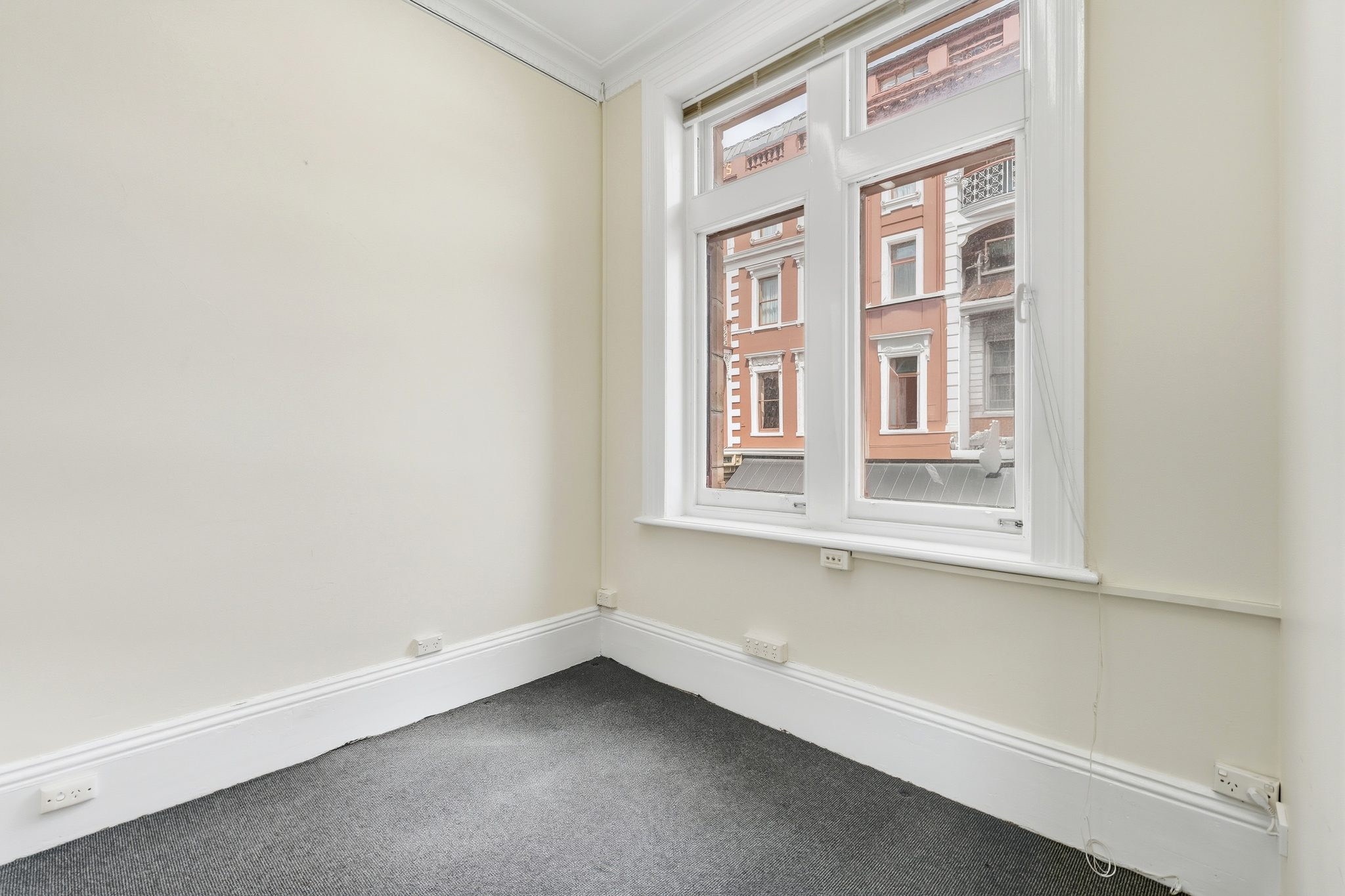 Corner of a carpeted room, sash window looking onto the painted facade of the building opposite.