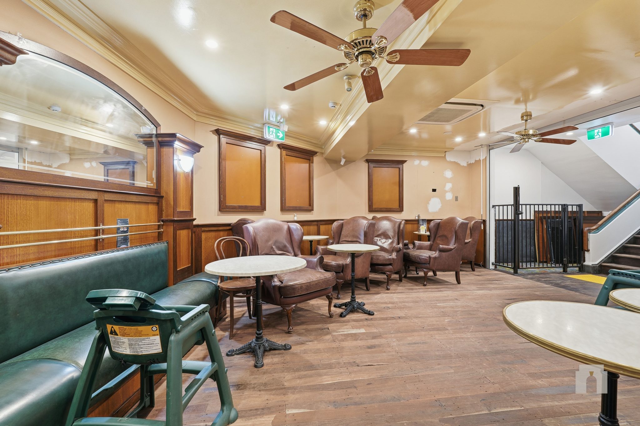 Leather wing chairs and cafe tables along a panelled wall, staircase behind railing at right.