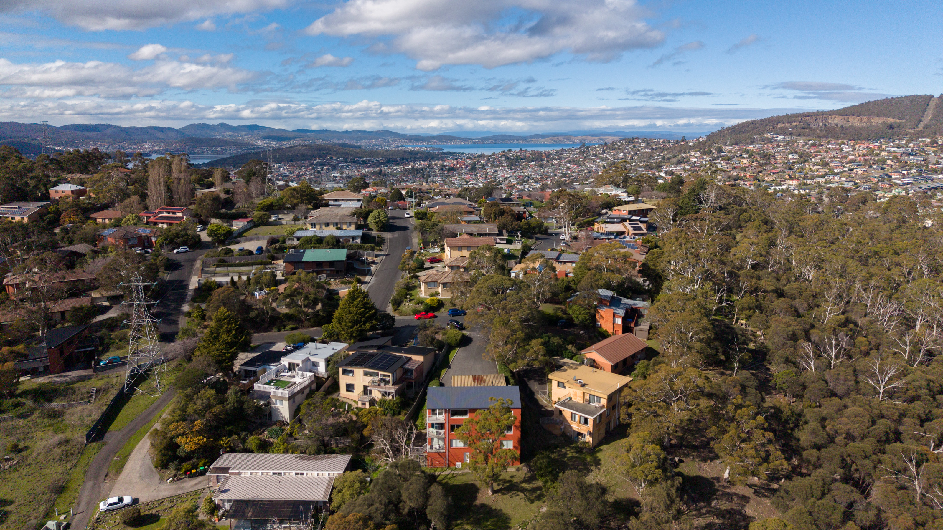 Aerial view over Lenah Valley rooftops and bushland, with the Derwent and the hills beyond.