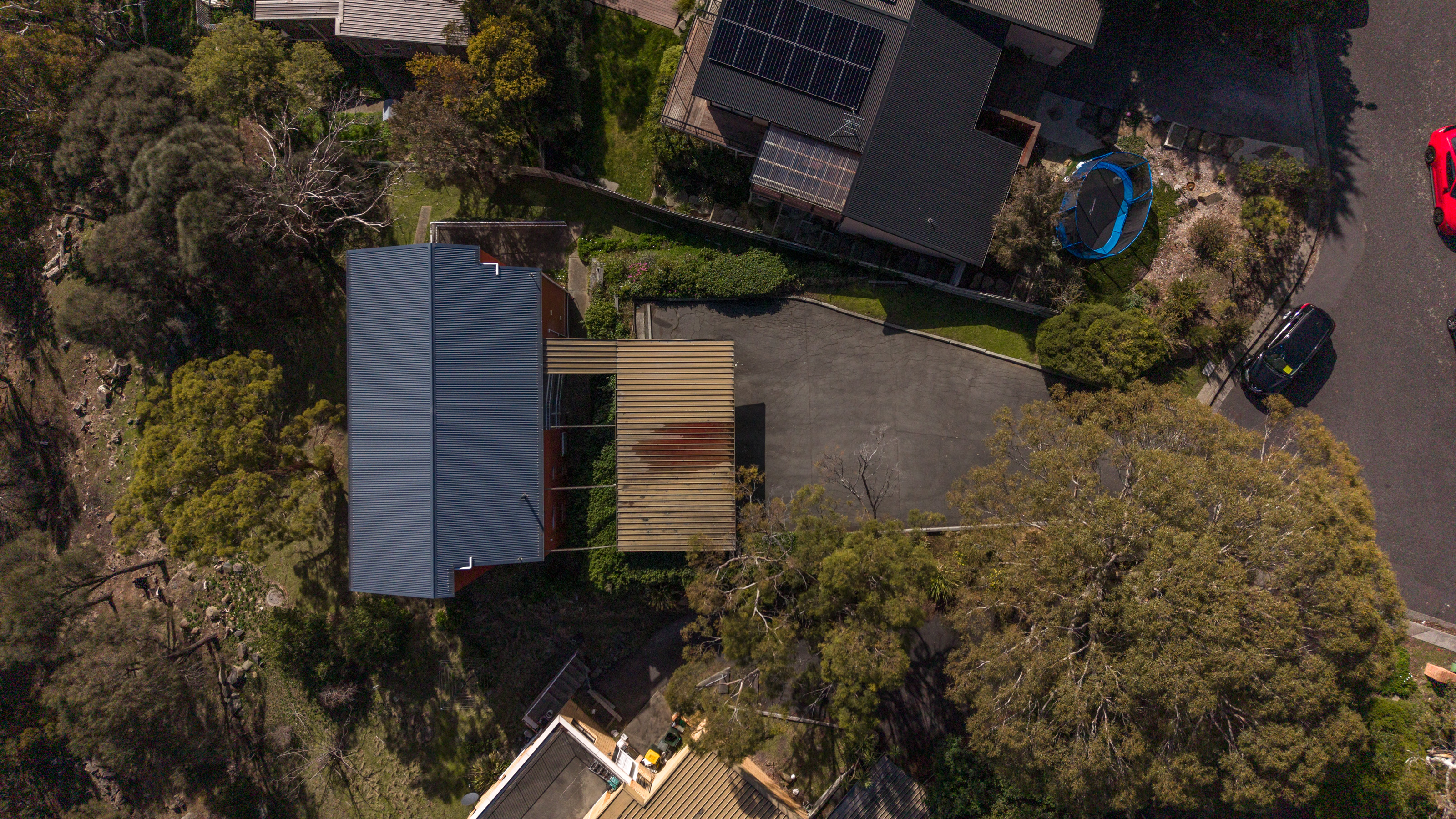 Aerial view looking straight down on the building, its ribbed metal roof running the length of the frame, with a slatted timber structure and the concrete parking area alongside and bushland dropping away to one side.