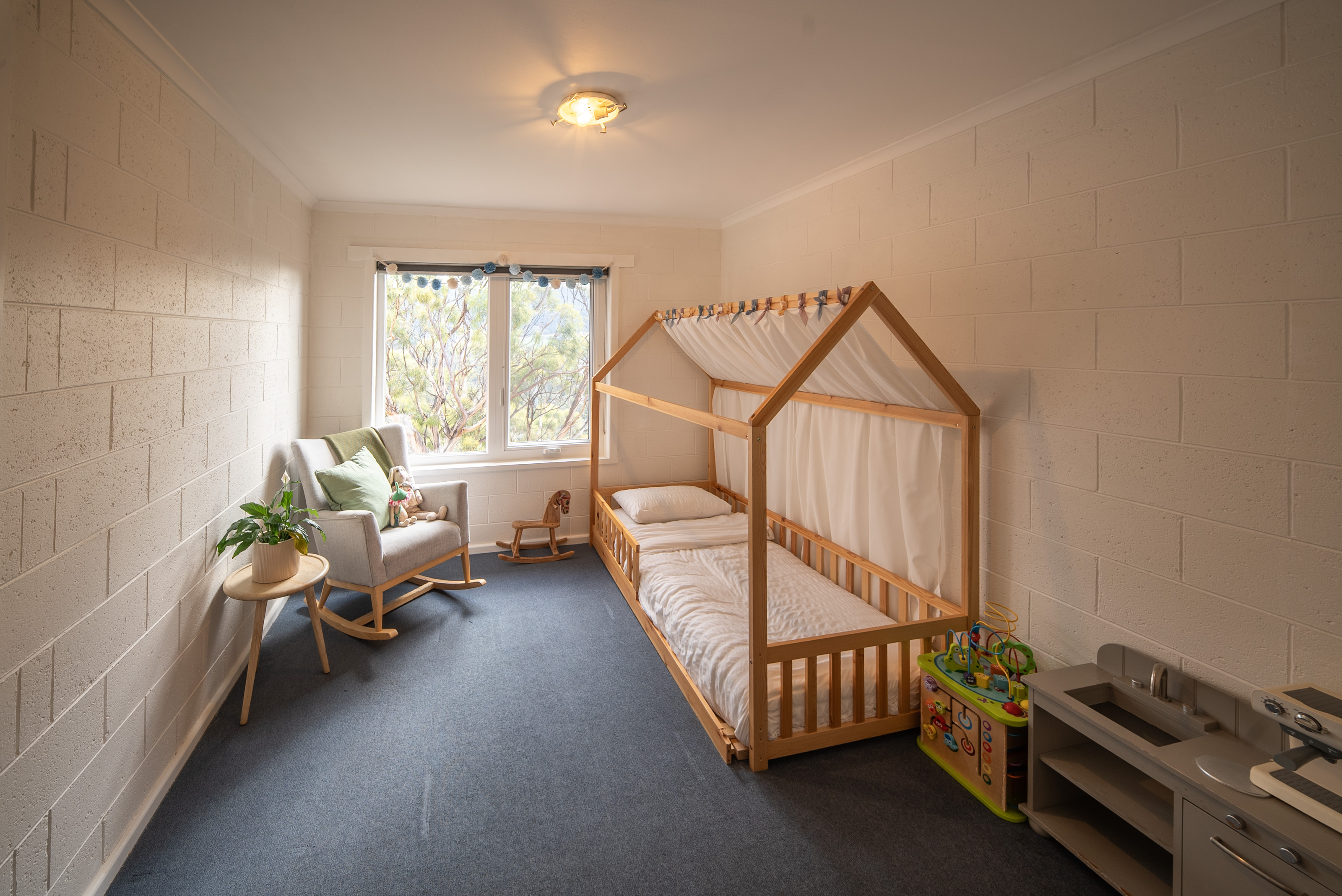 Second bedroom set up as a child's room, with a timber house frame bed, a rocking chair by the window and grey carpet.