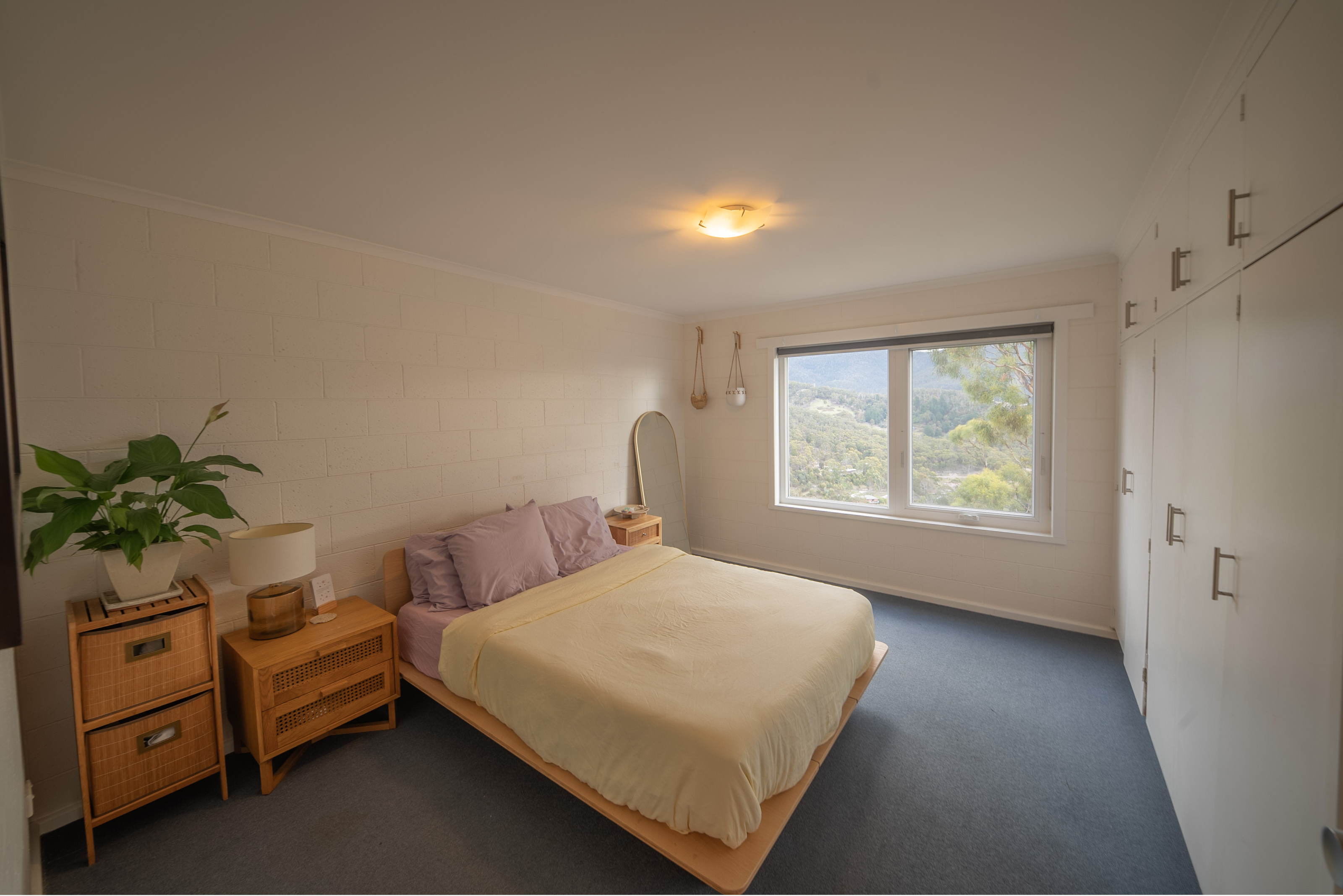 Main bedroom with a double bed, built in white wardrobes along one wall, and a window looking out to bush and hills.