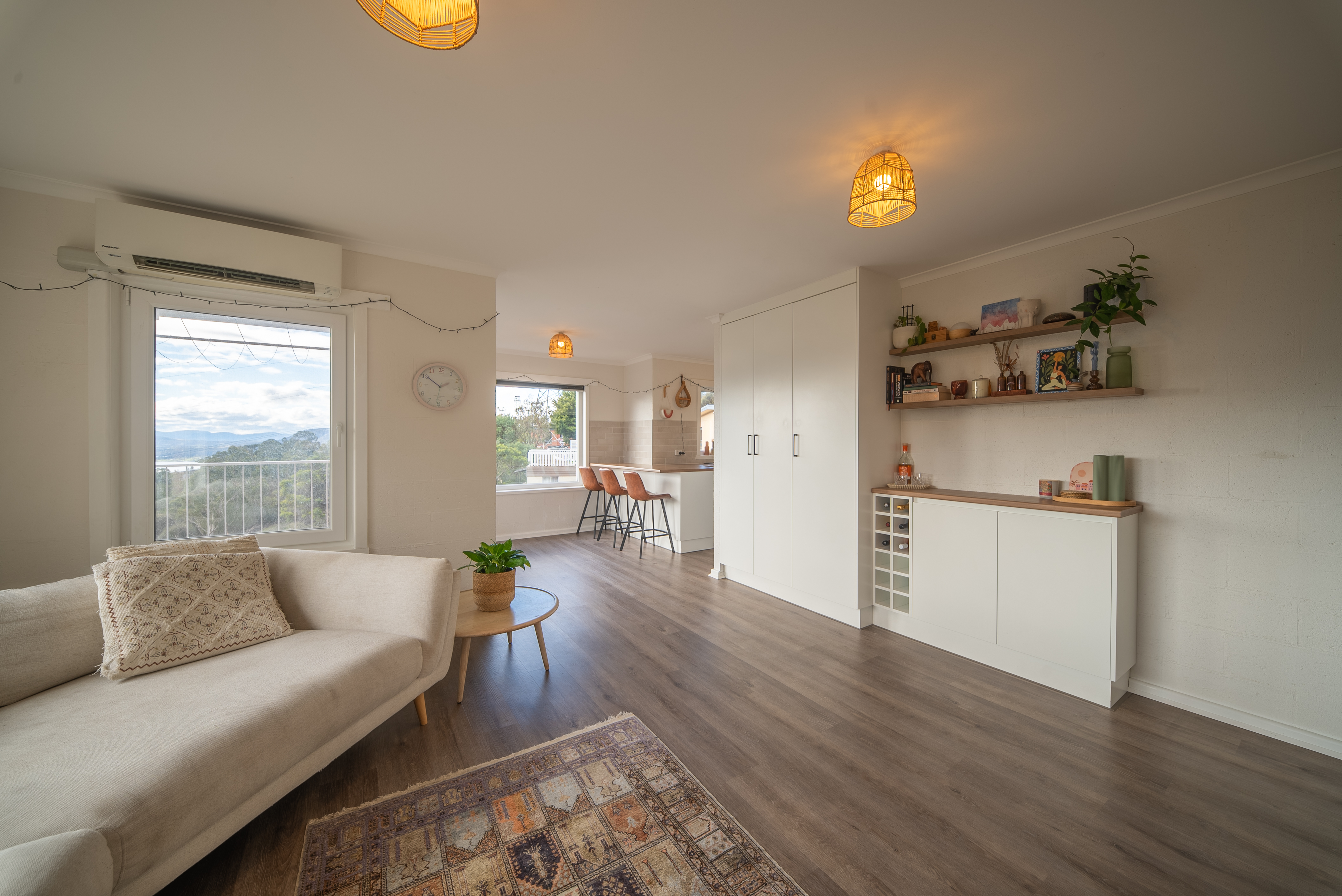Living room looking through to the kitchen, with a breakfast bar and stools, white cabinetry and open timber shelves.