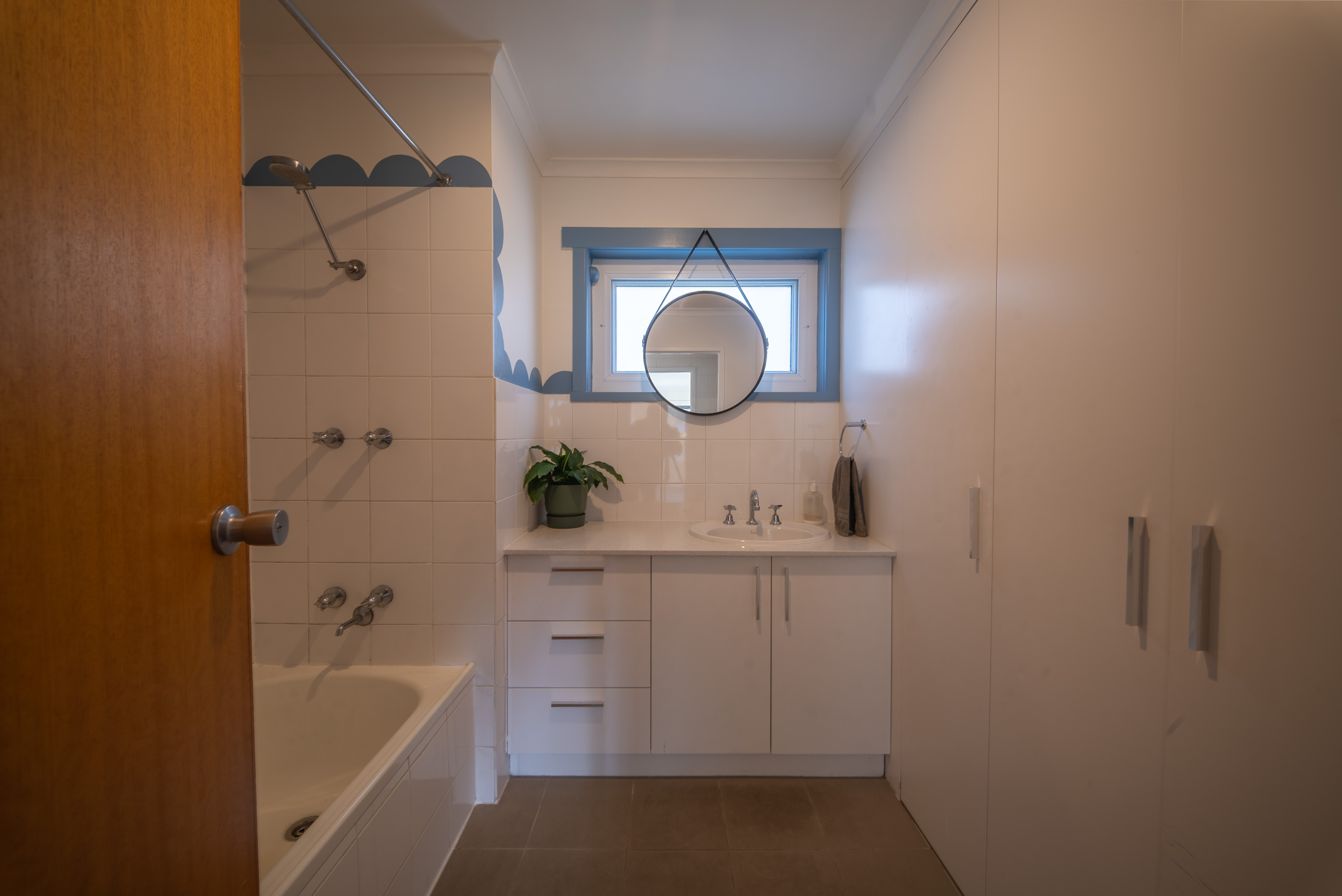 Bathroom with a bath and shower over, a white vanity, a round mirror and a blue framed window.
