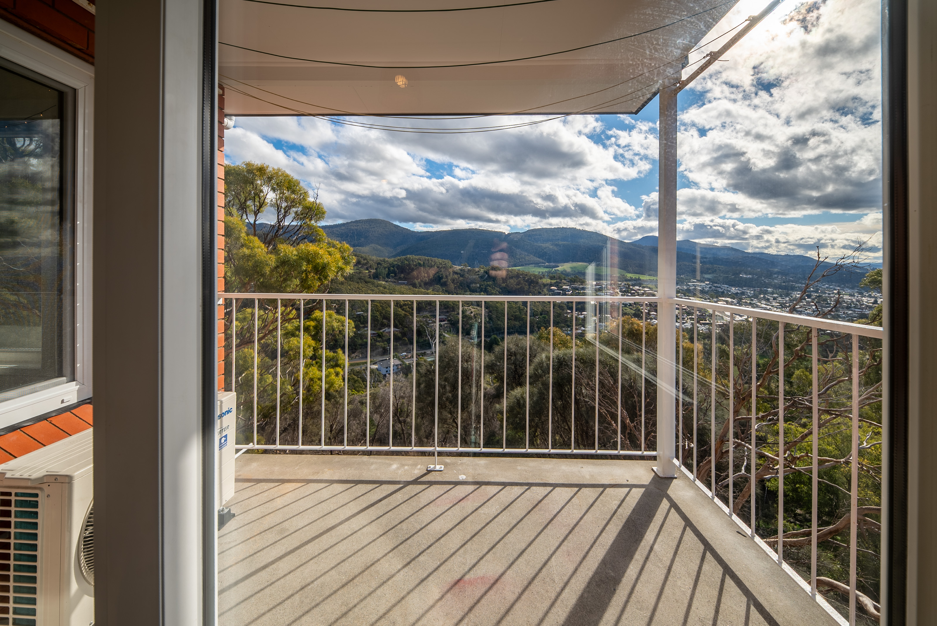 The balcony, with a white railing, a shade sail overhead and a view across bushland to the hills.