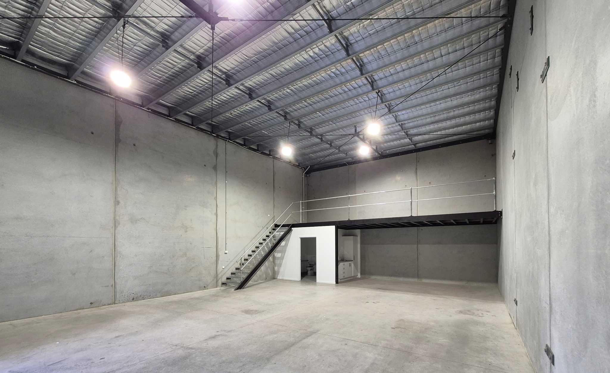Warehouse floor with concrete panel walls, high-bay lights, and a stair up to the mezzanine.
