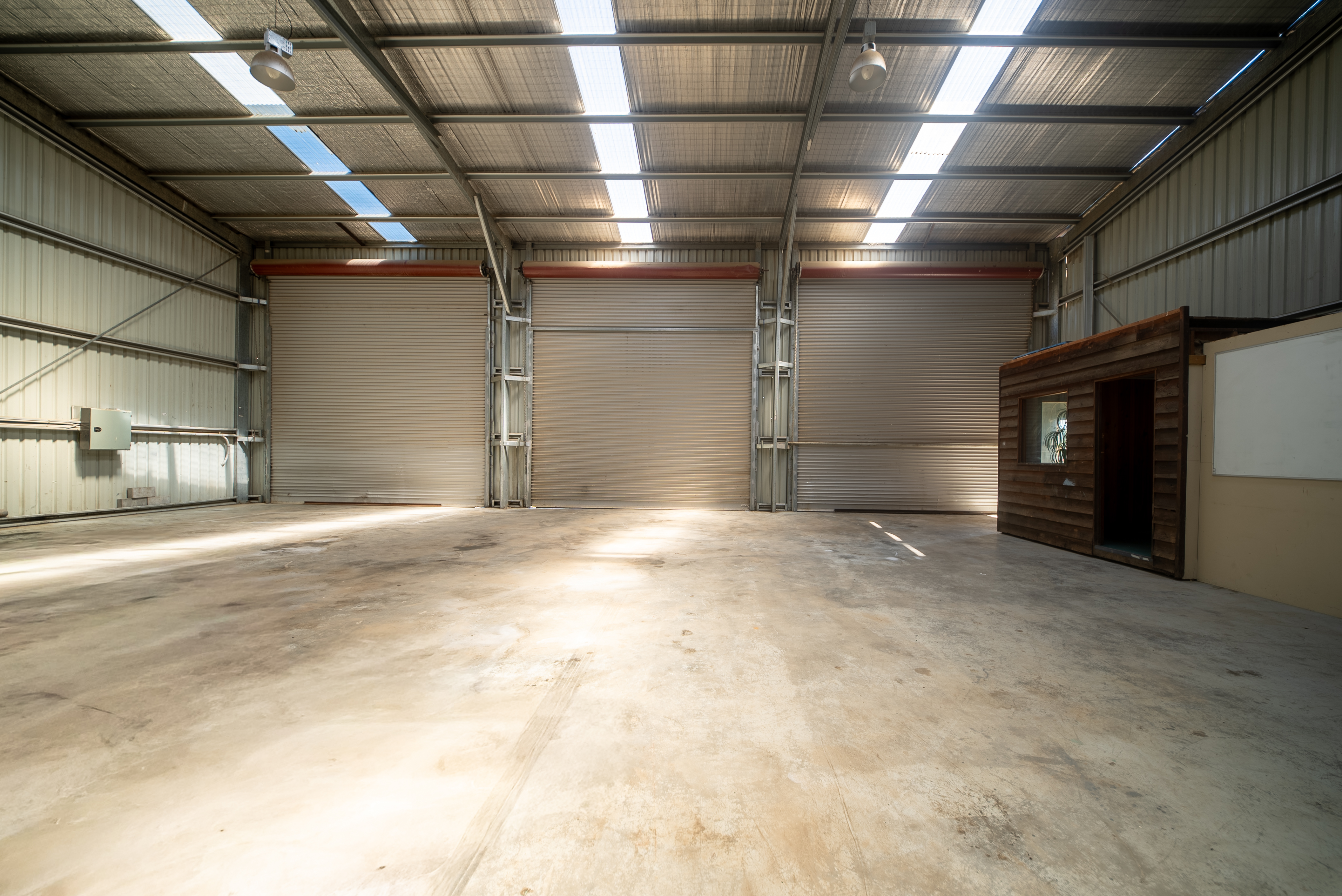Inside the warehouse looking back at the three roller doors, with clear roof sheets, high-bay lights and a concrete floor.