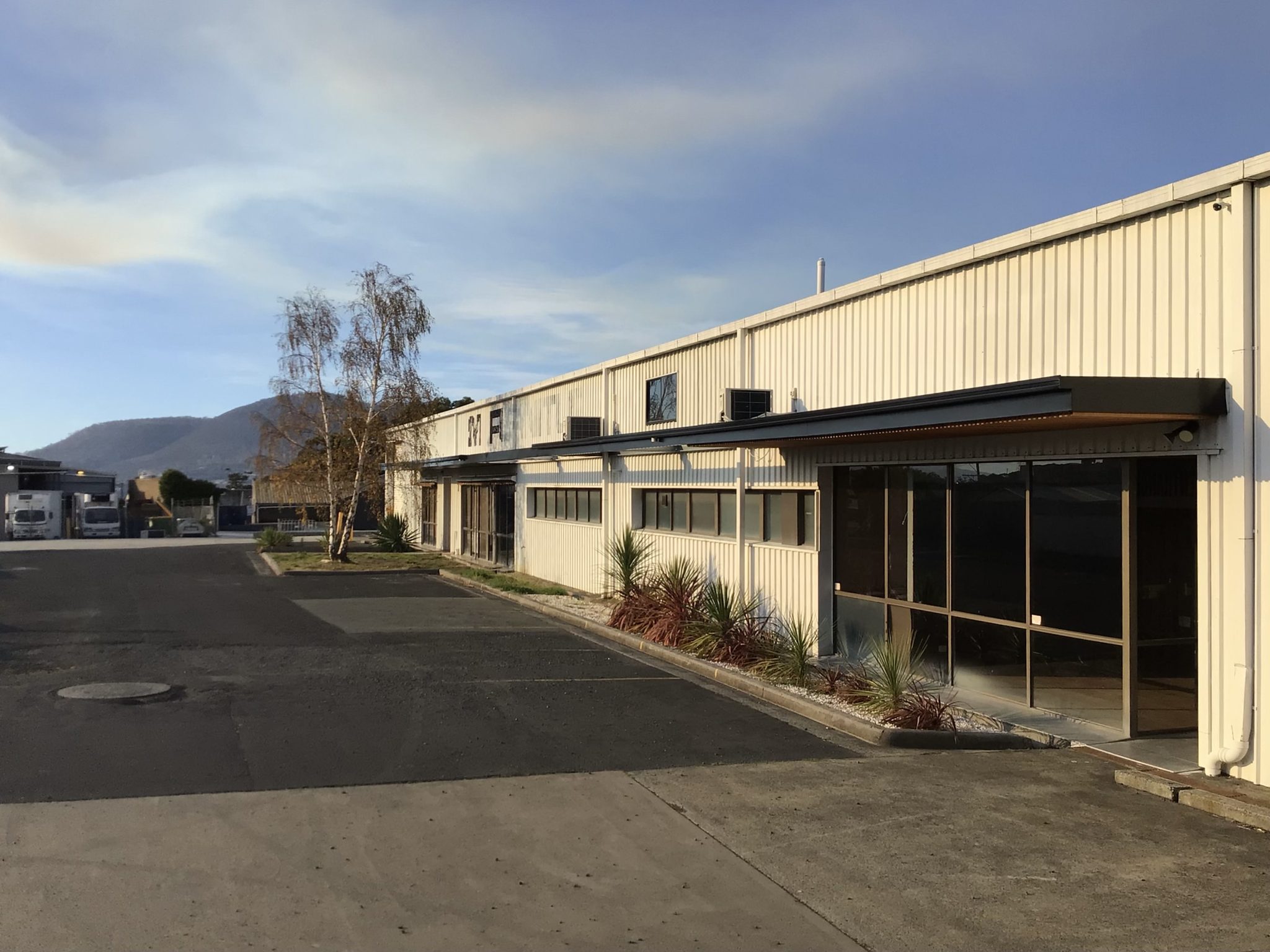 Side of the building with a glazed office entry under an awning, sealed driveway and hills behind.