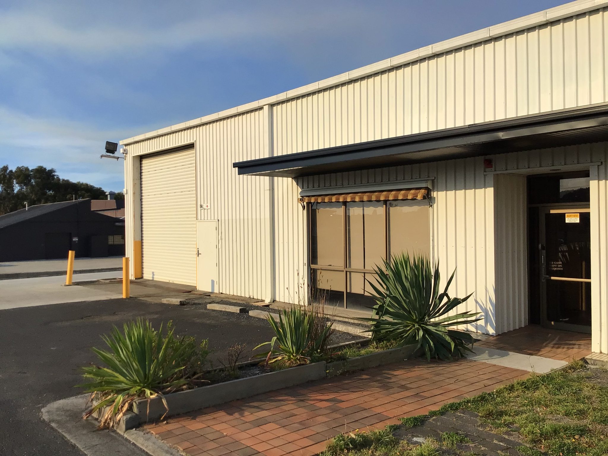 Further roller shutter door along the building, with an awning over an office window and a paved path.